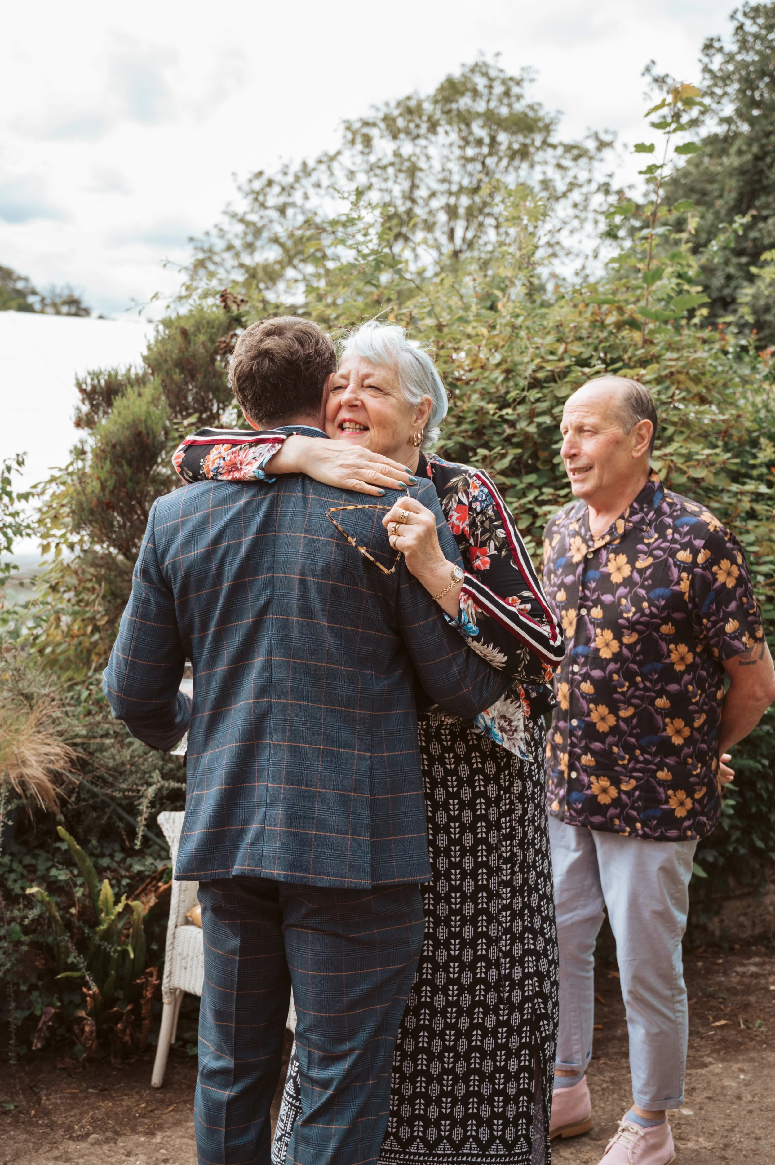Three people at an outdoor gathering, with two of them hugging, surrounded by greenery.