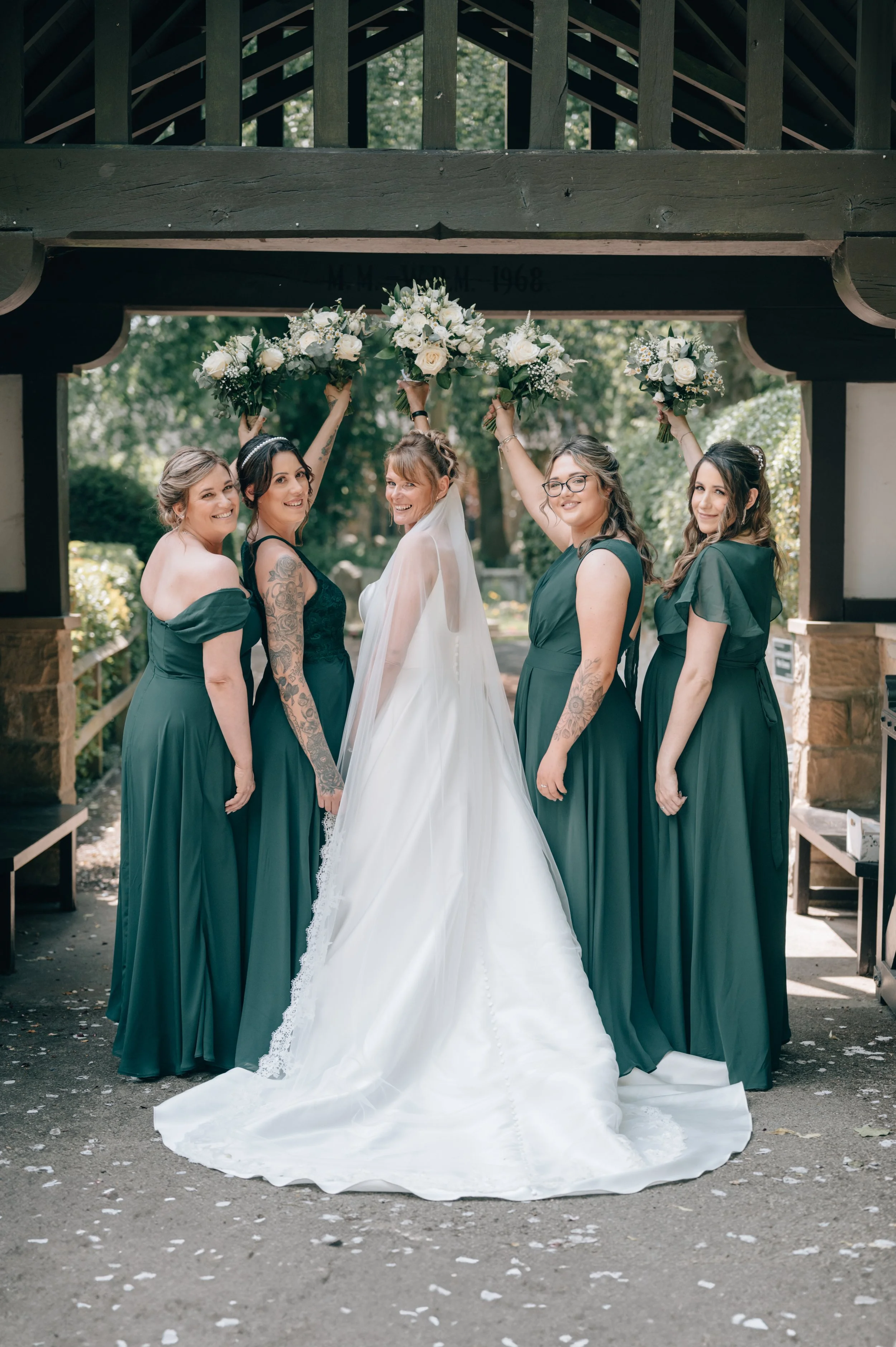 A bride in a white wedding dress and veil stands with four bridesmaids in green dresses, holding bouquets of white flowers overhead, under a wooden archway outdoors.