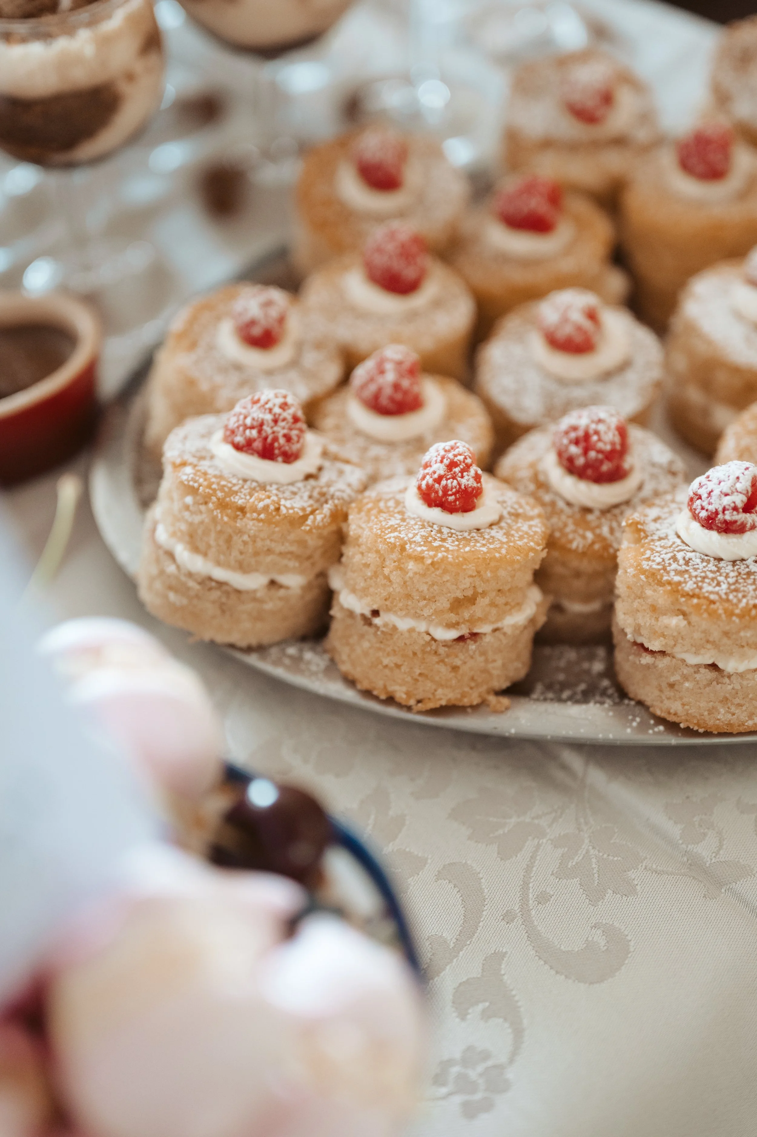 Plate of mini cakes topped with raspberries and powdered sugar, with a blurred dessert in the foreground and additional dessert items in the background.