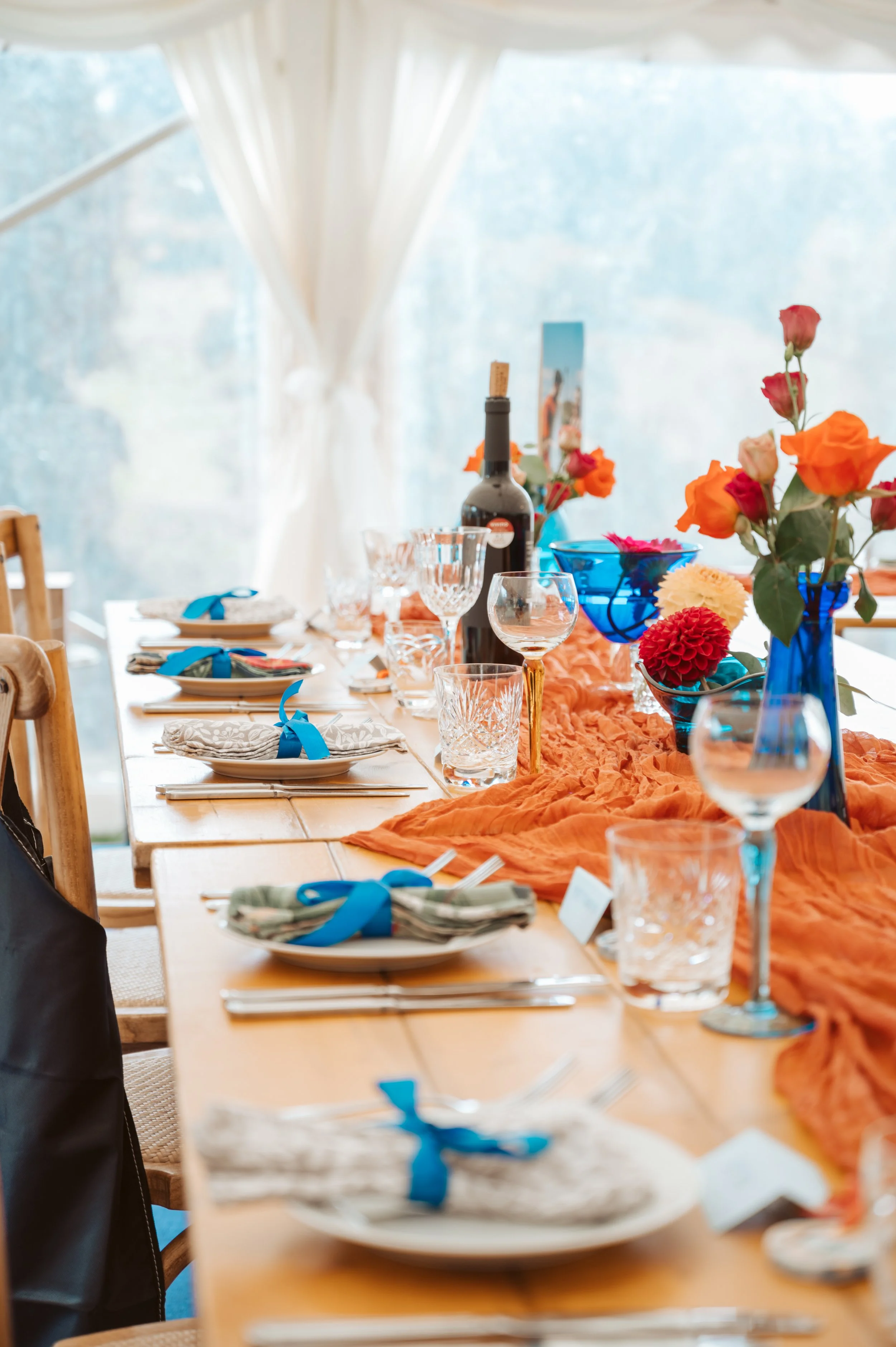 A decorated dining table with a table runner, colorful flowers in vases, wine glasses, cutlery, and napkins tied with blue ribbons, set in a bright room with large windows and white curtains.