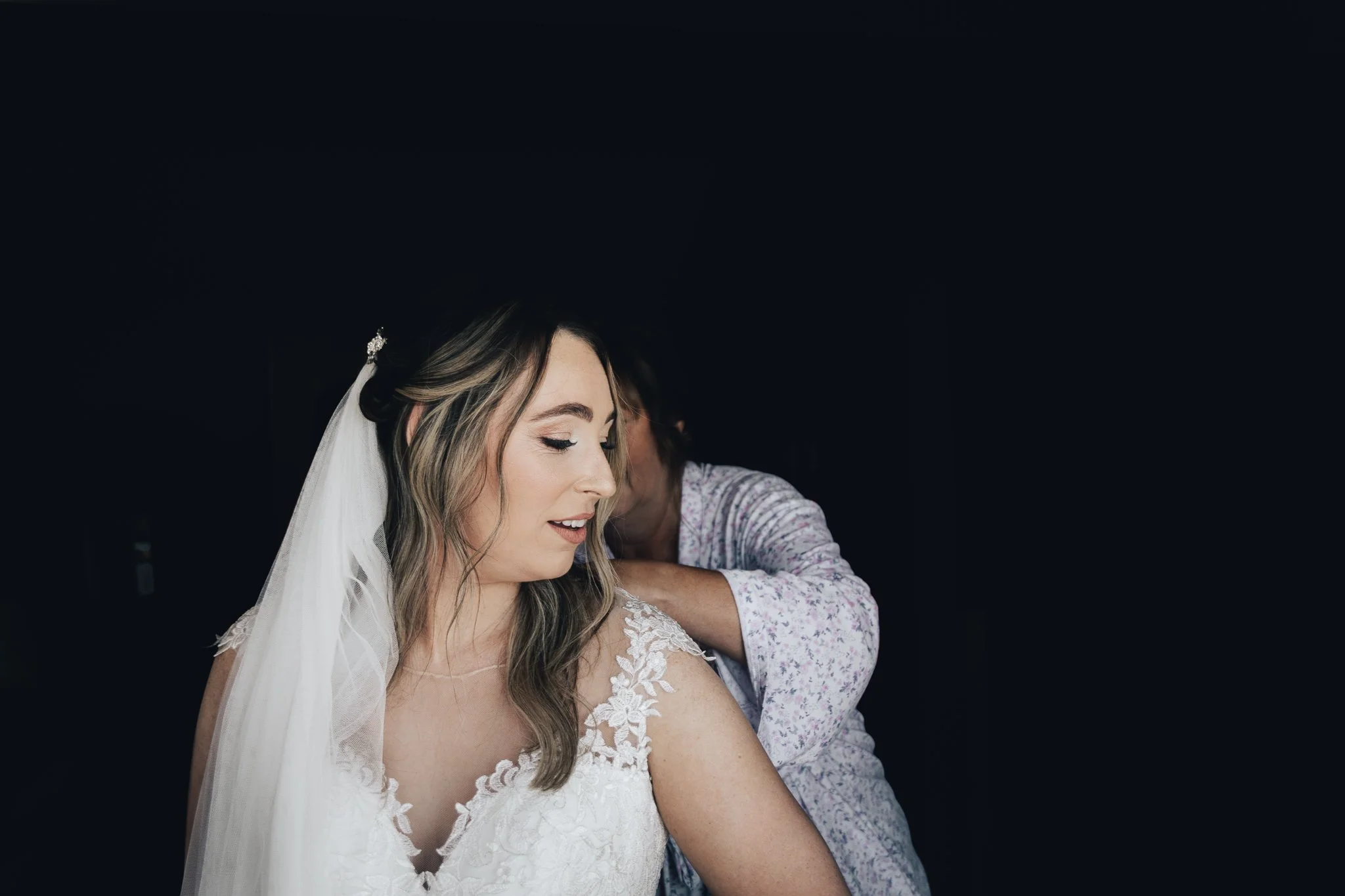 A bride in a white lace dress has her wedding veil adjusted by an older woman, possibly her mother, against a dark background.