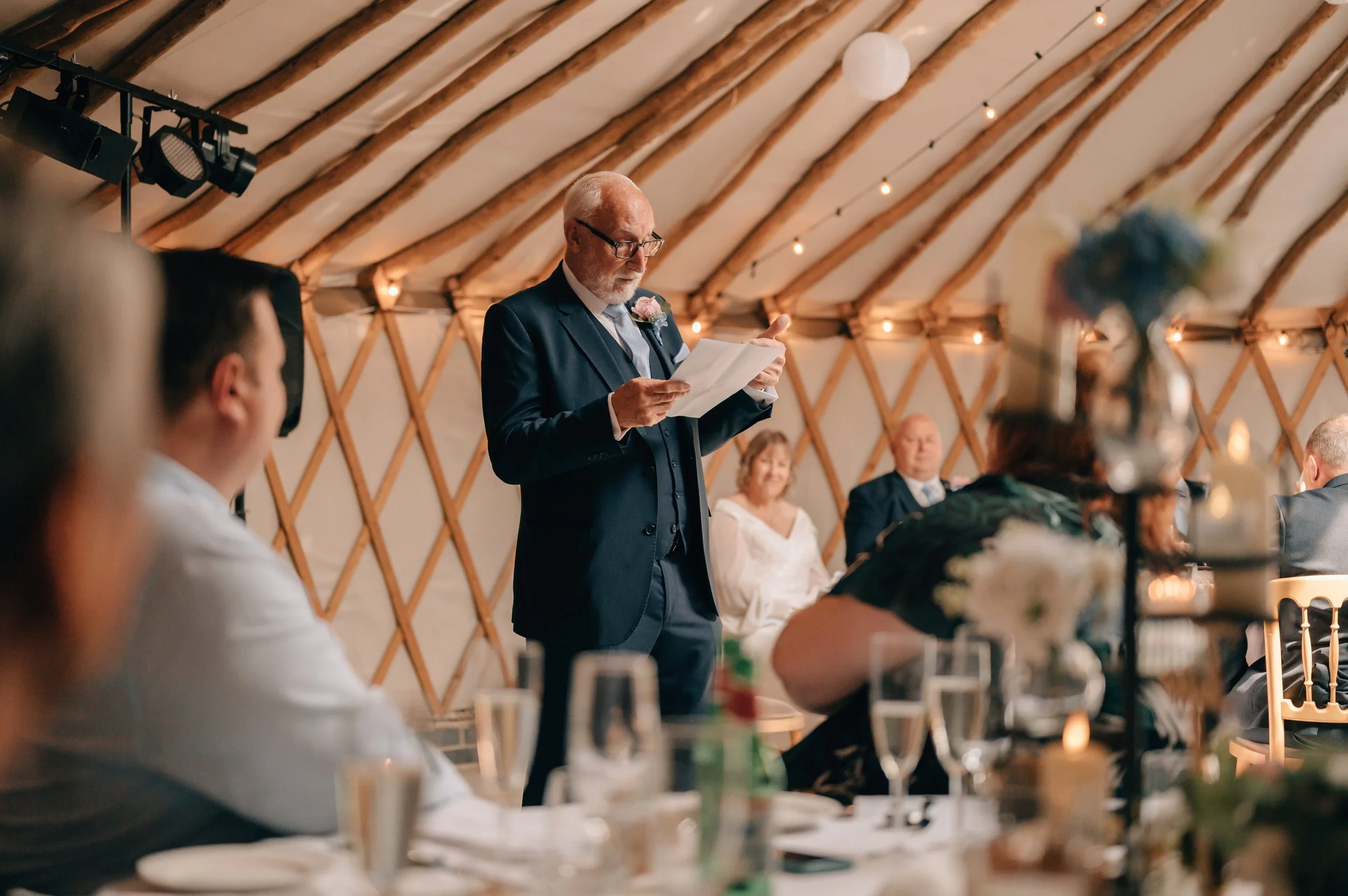 An elderly man in a suit and glasses giving a speech at a wedding reception, standing in front of seated guests inside a decorated venue with wooden beams and string lights.