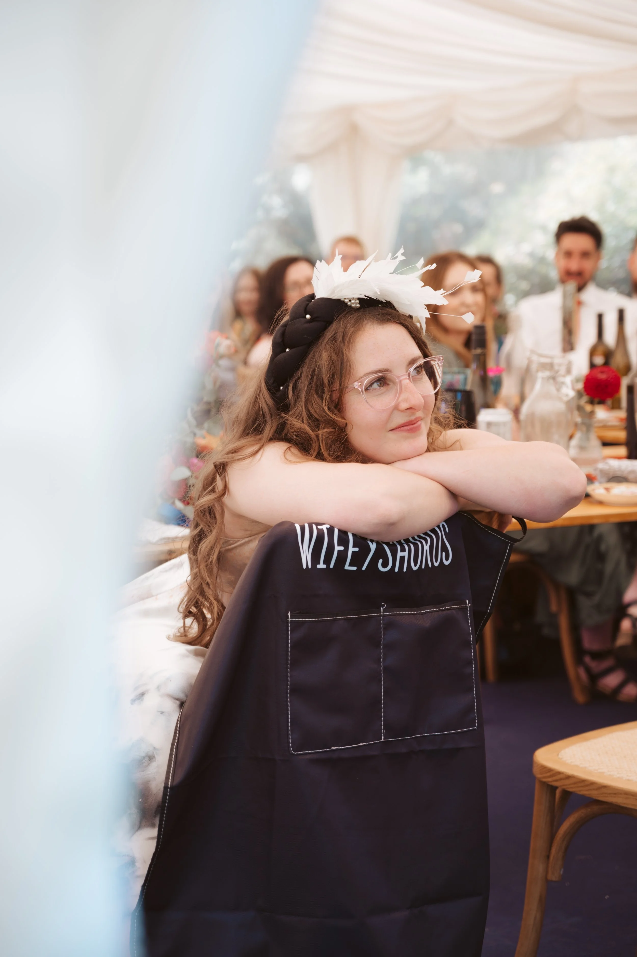 A woman with curly hair and glasses resting her head on her crossed arms on a black tote bag at a table during a celebration, with friends and family in the background in a decorated tent.
