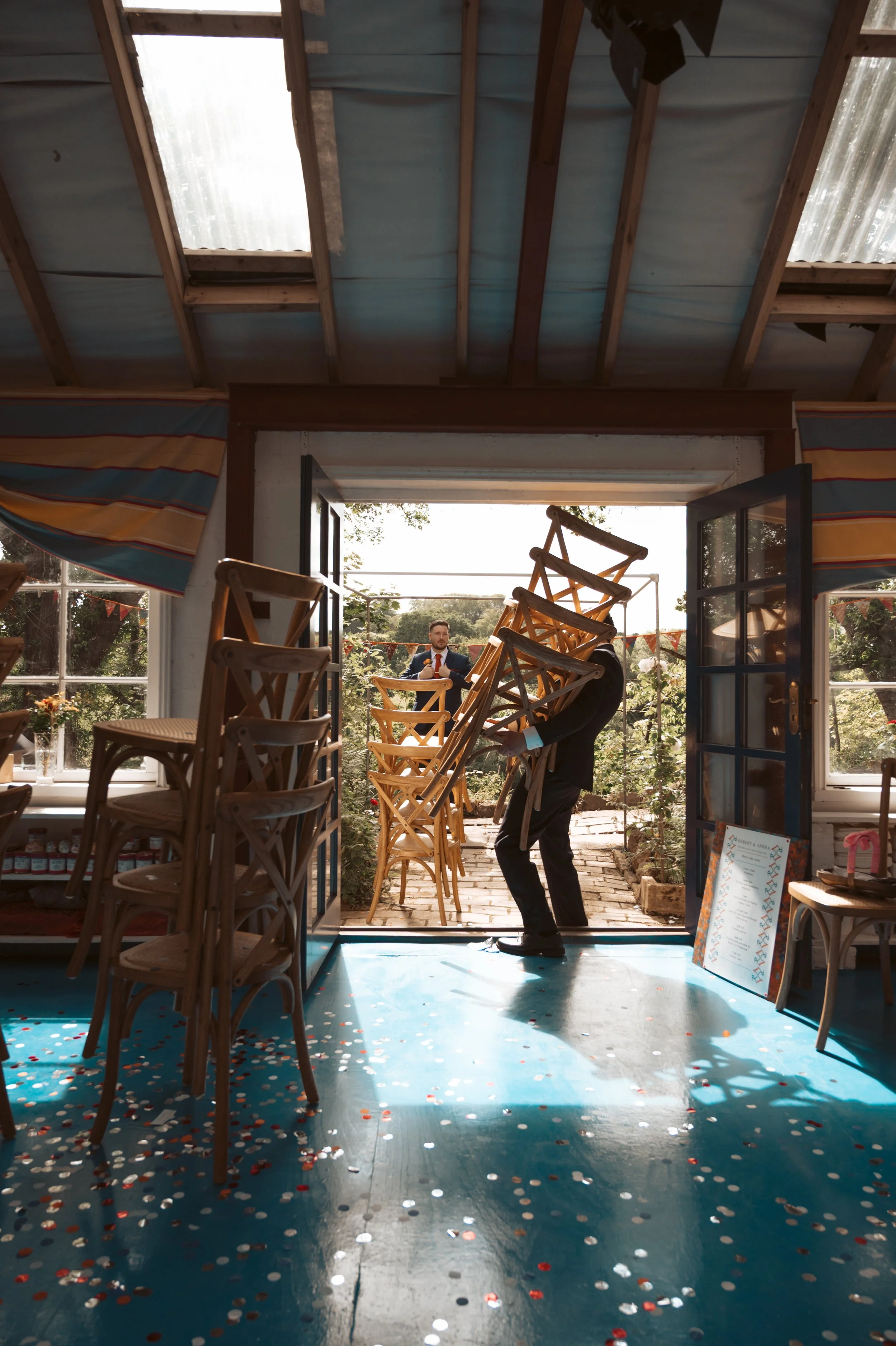 A person carrying multiple wooden chairs into a decorated venue with confetti on the floor, with two men in the background outside near a garden.