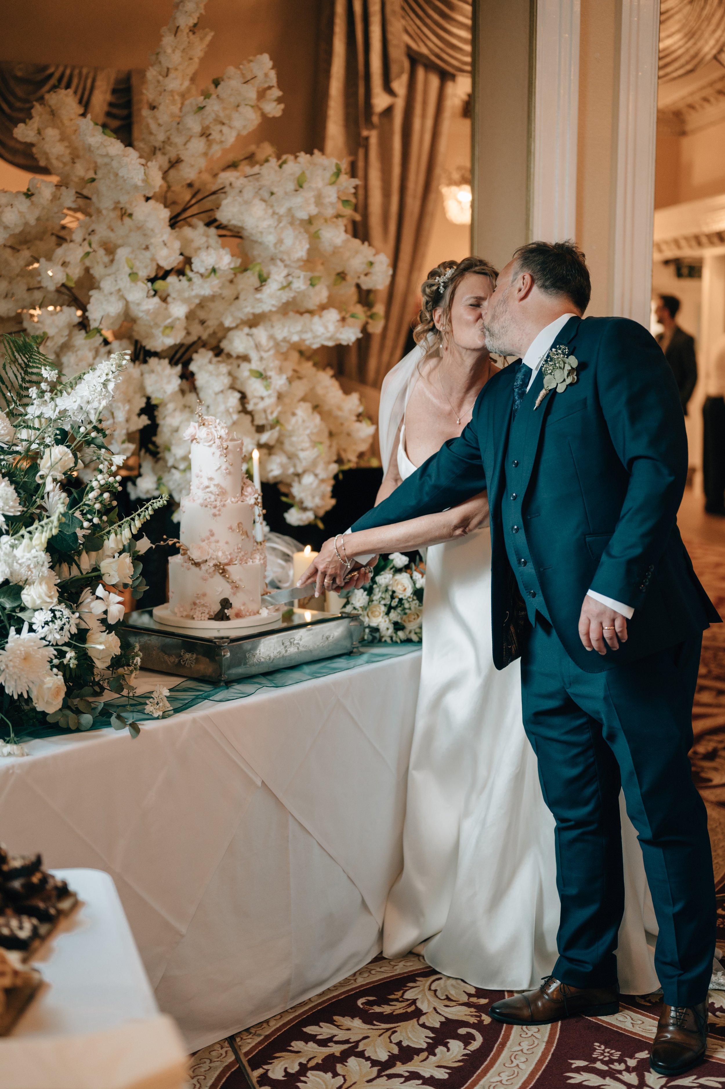 A bride and groom share a kiss while cutting a wedding cake at their reception. The table is decorated with white flowers and a large floral arrangement in the background.
