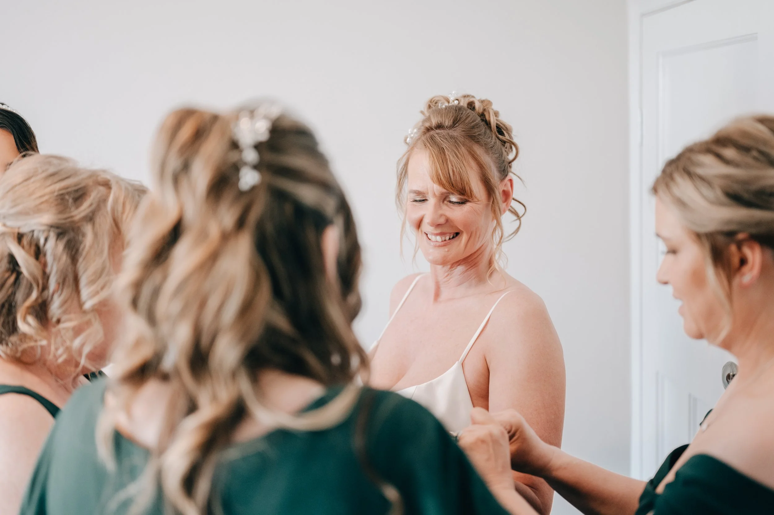 A woman with red hair in a dress is smiling with eyes closed as she is surrounded by women helping her get ready, possibly for a wedding.