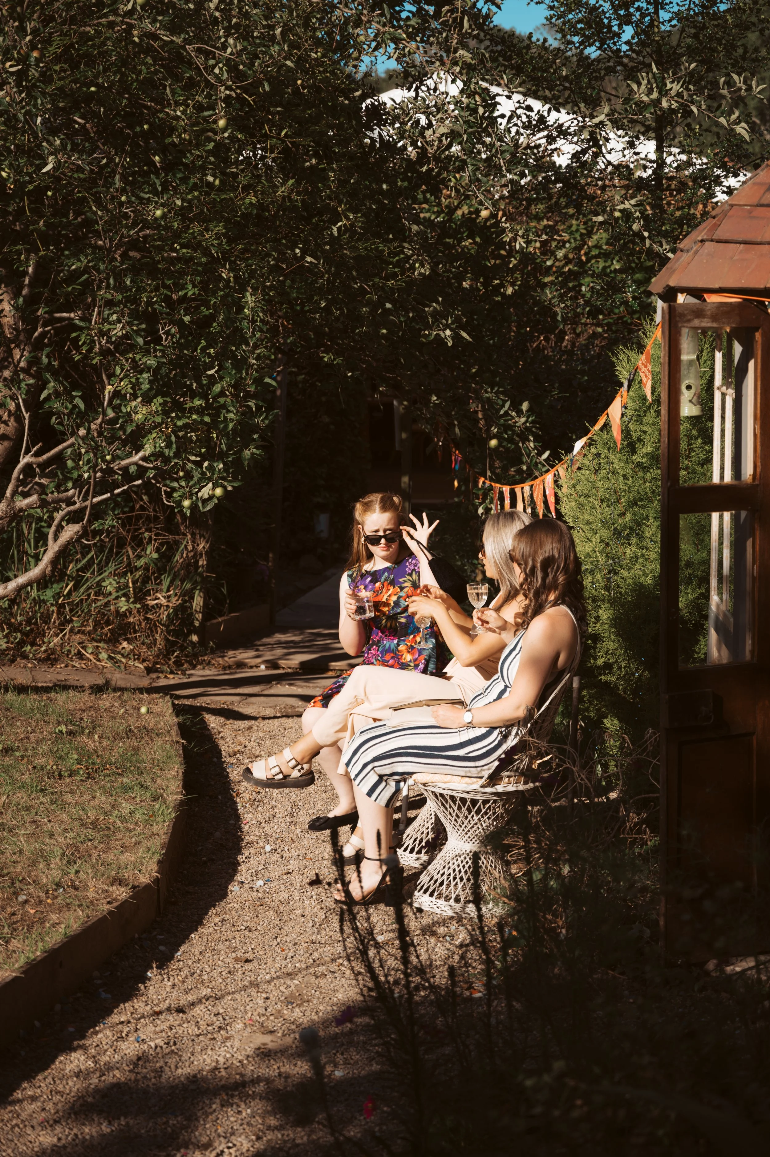 Three women sitting on chairs outdoors, enjoying drinks, in a garden setting with trees and decorated with strings of triangular flags.