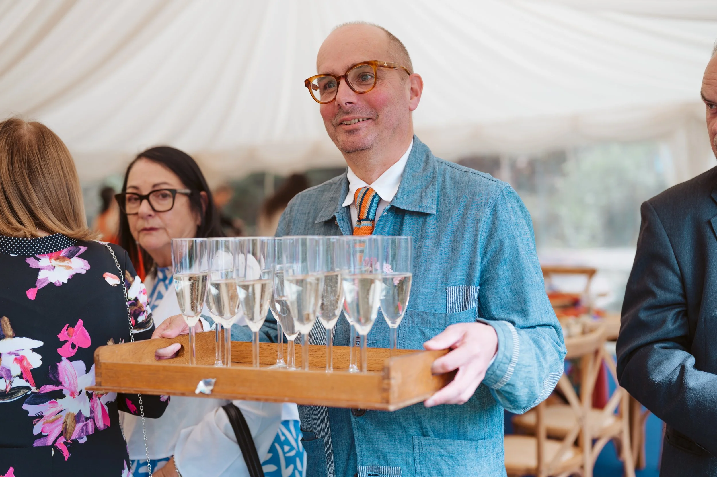 A man in glasses and a blue jacket holding a tray with several glasses of champagne at a social event in a tented venue.