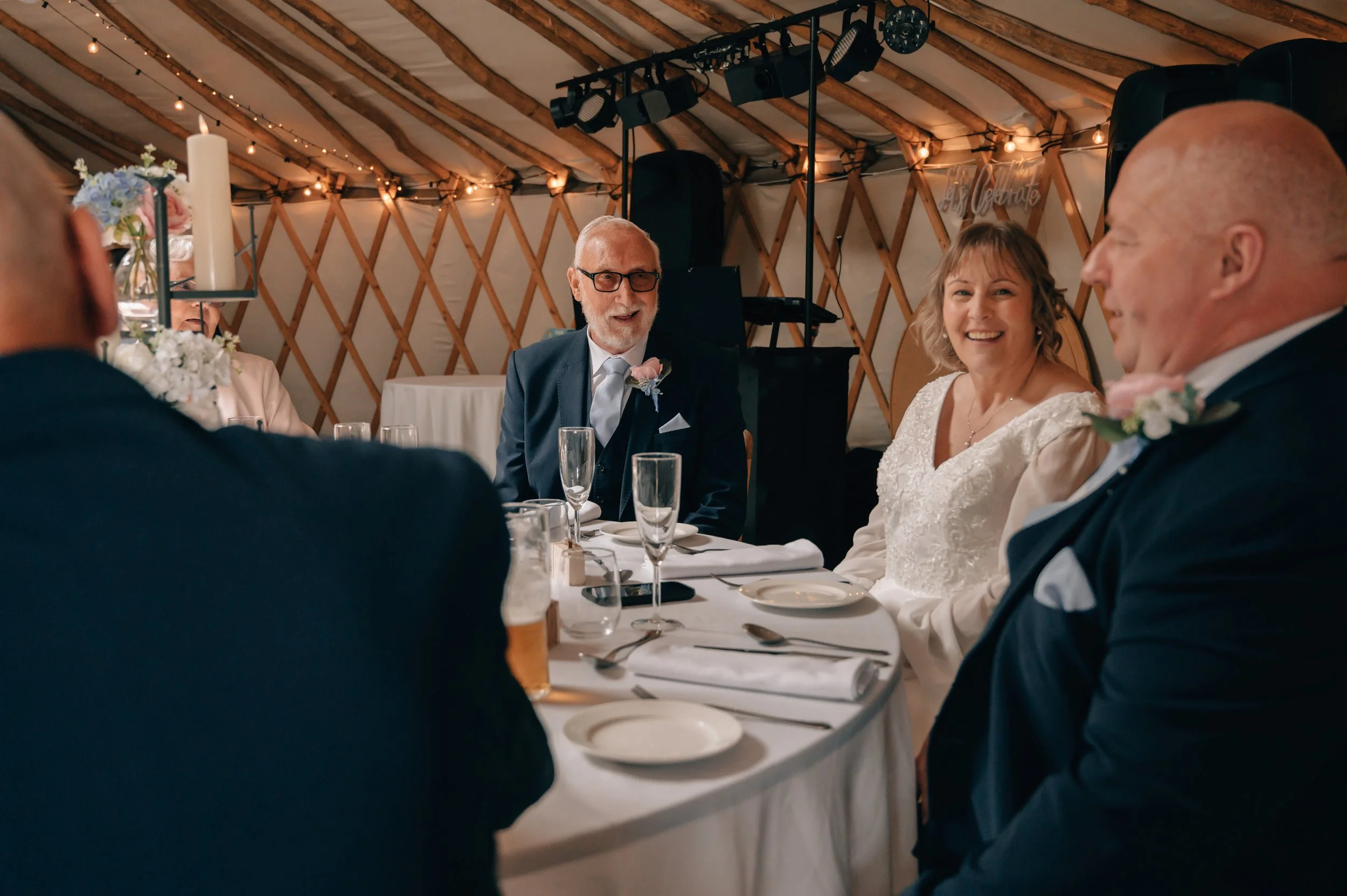 People sitting at a round table during a wedding reception in a decorated tent, smiling and talking, with glasses and plates on the table.