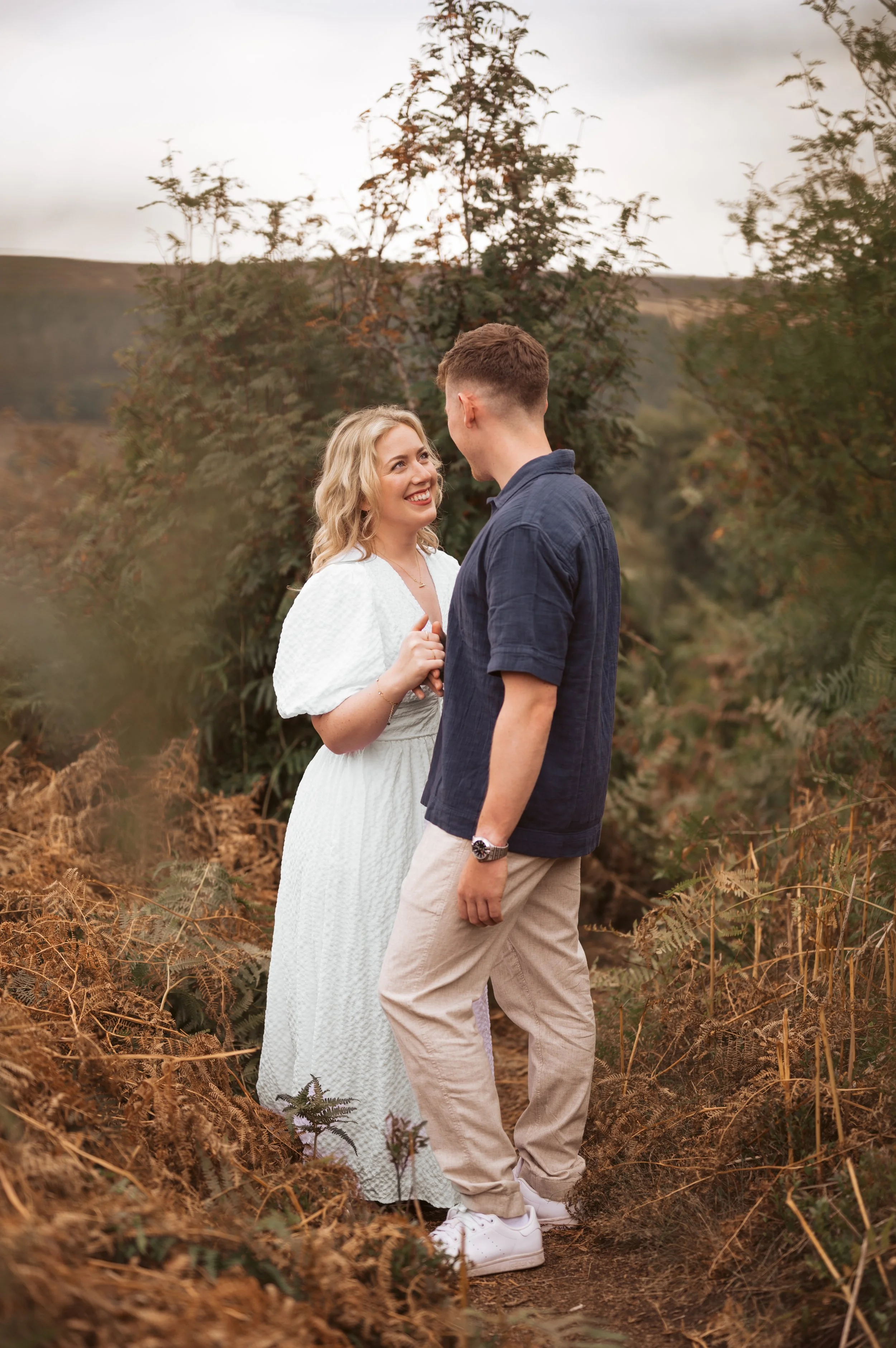 A couple standing on a dirt path outdoors, facing each other and smiling, surrounded by greenery and trees with a cloudy sky.