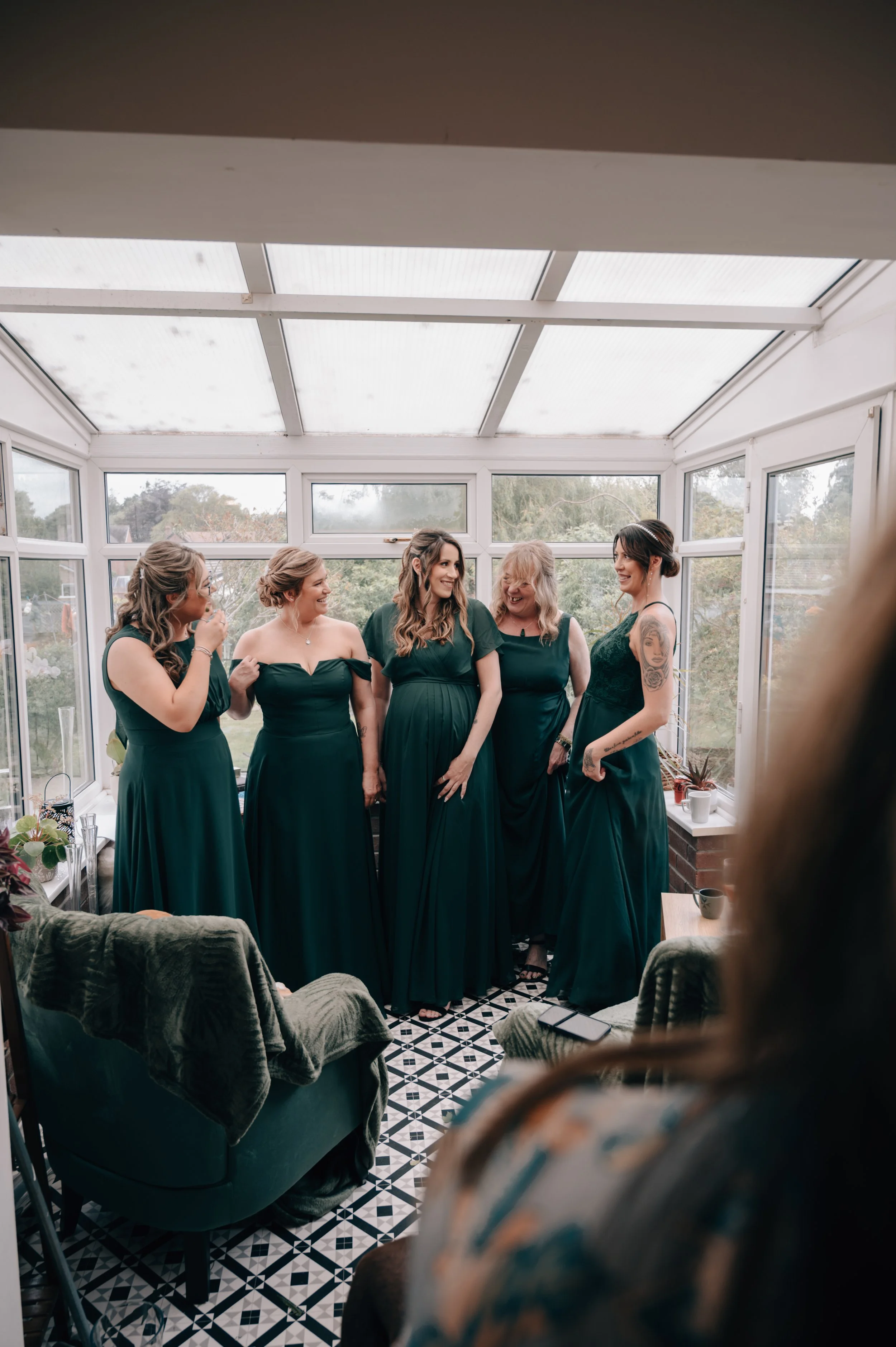 Group of five women dressed in dark green dresses in a sunroom, smiling and talking, with a woman partially visible in the foreground.