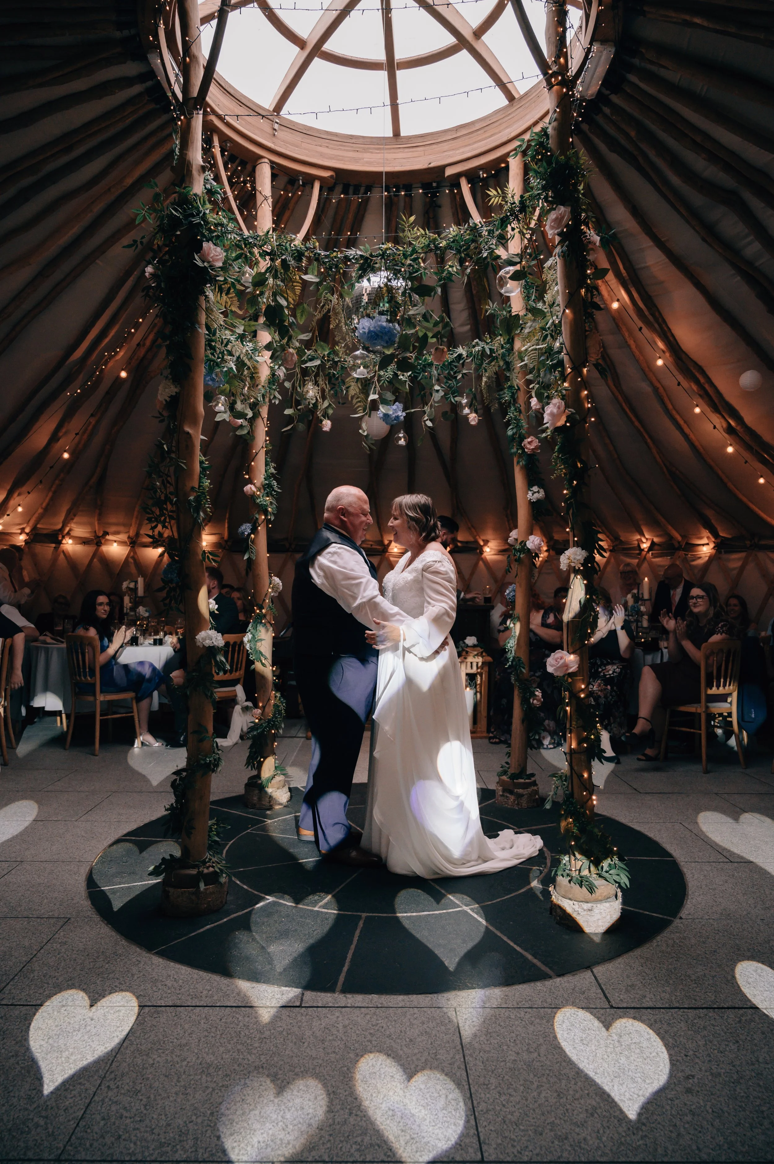 A bride and groom share a dance during their wedding reception inside a decorated tent with fairy lights and hanging greenery, surrounded by seated guests.