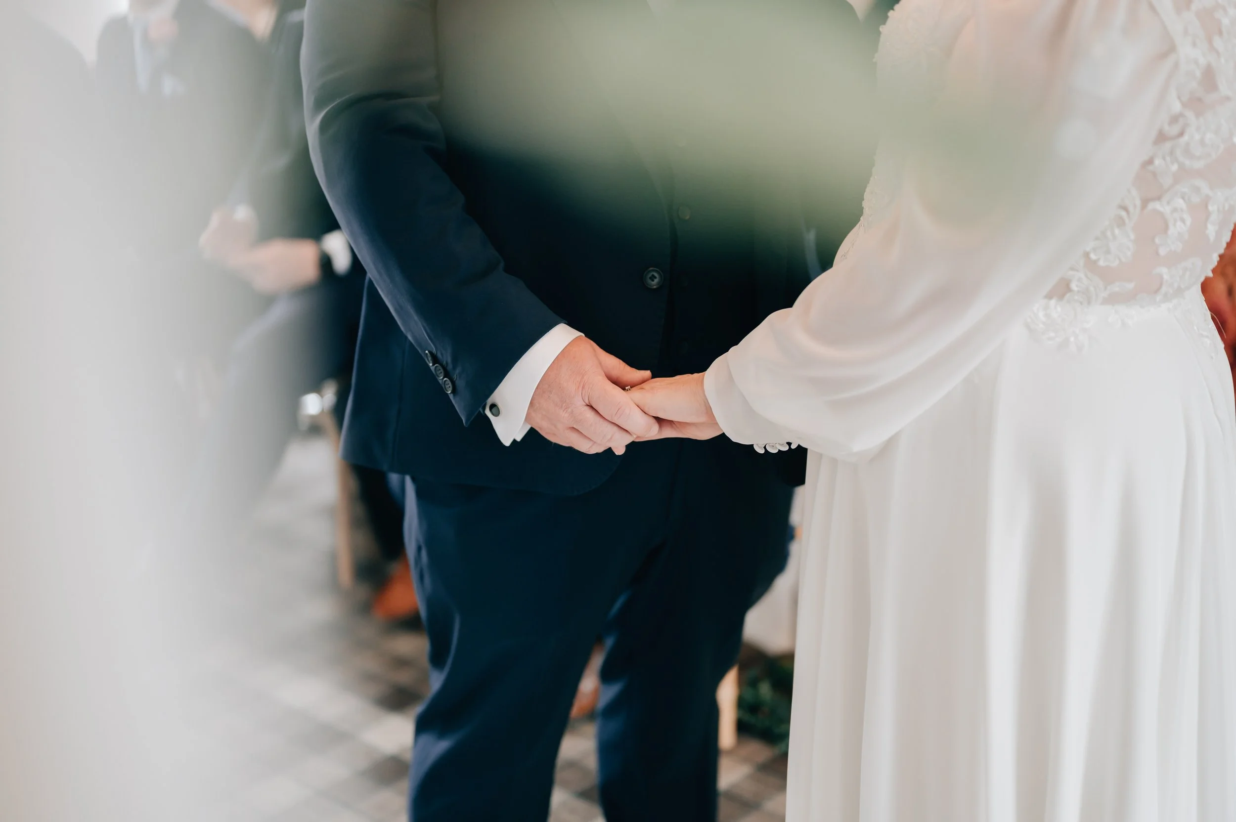 Close-up of a bride and groom holding hands during their wedding ceremony, with the bride in a white dress with lace details and the groom in a dark suit.