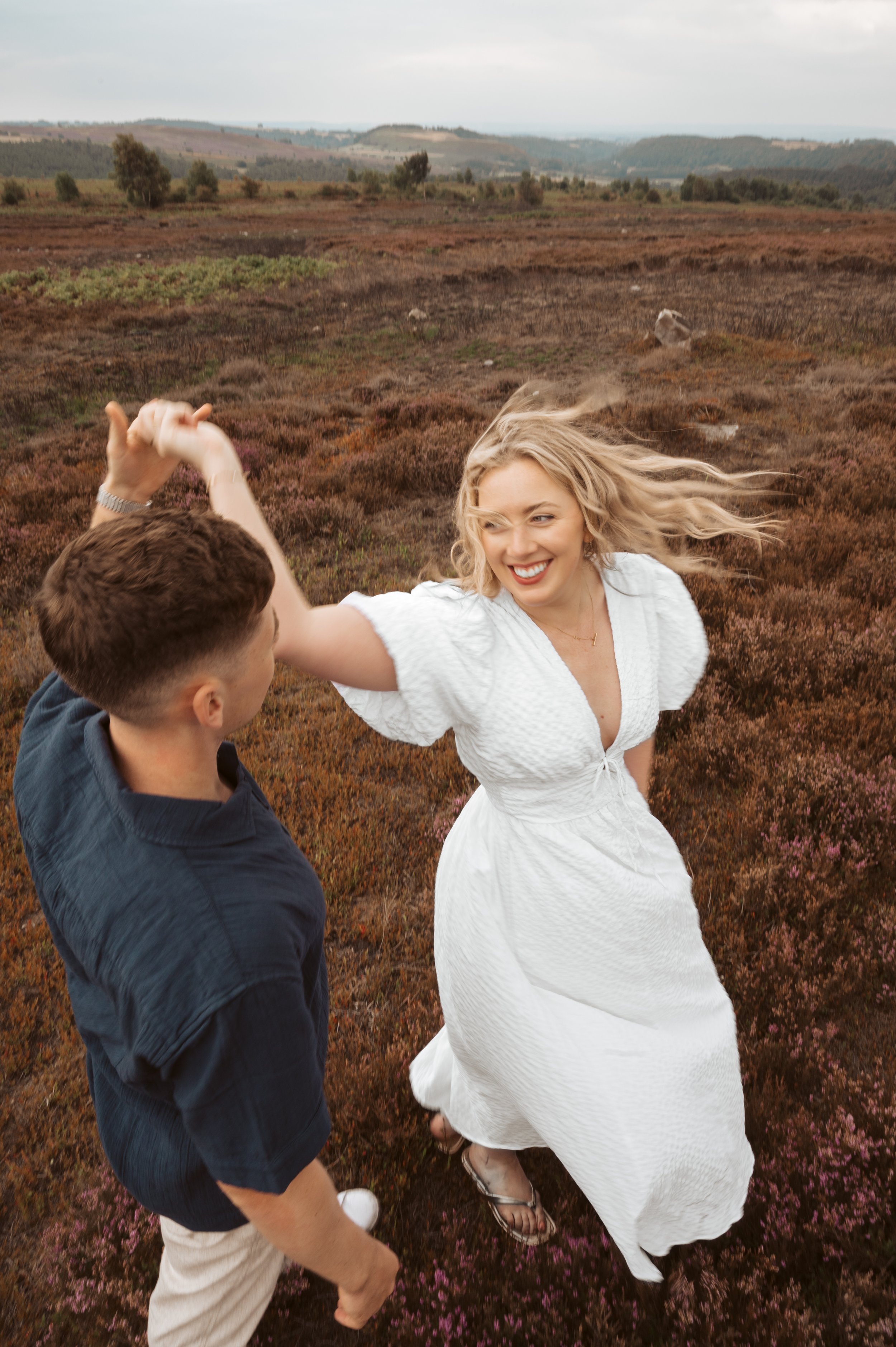 A woman in a white dress dancing with a man in a dark shirt in a scenic field with rolling hills in the background.