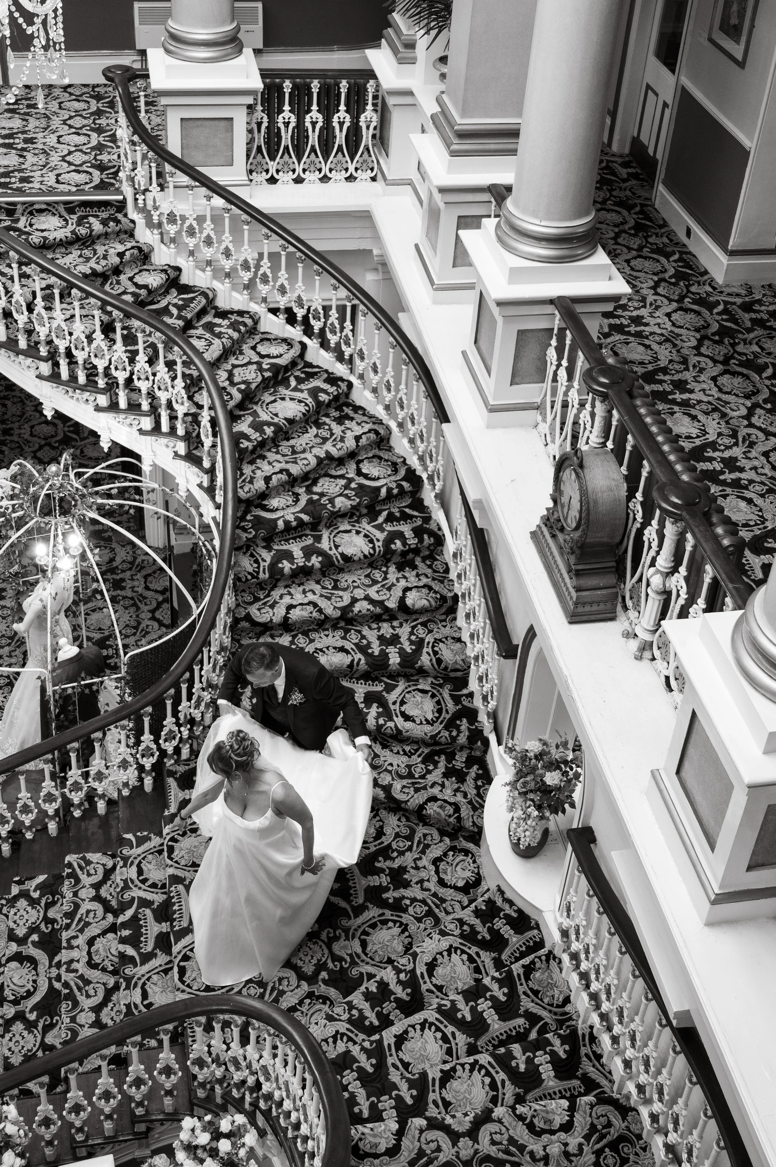 A bride and groom' dancing on a grand, curved staircase decorated with floral-patterned carpet. The staircase has ornate white and black railings, with a decorative clock on the wall nearby.