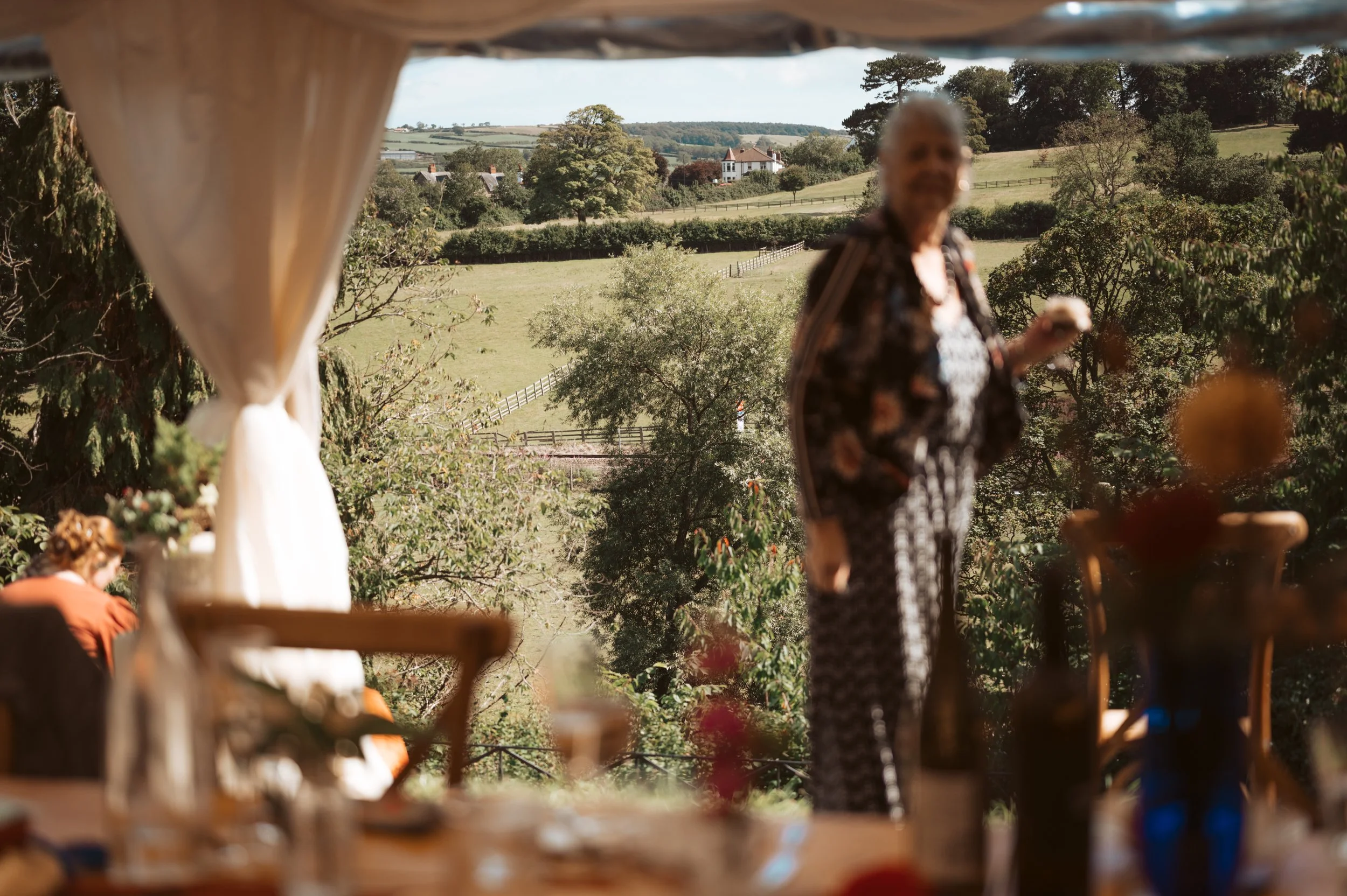 Blurry image of a woman standing outdoors during daytime with a scenic rural landscape in the background, seen through an opening with curtains.