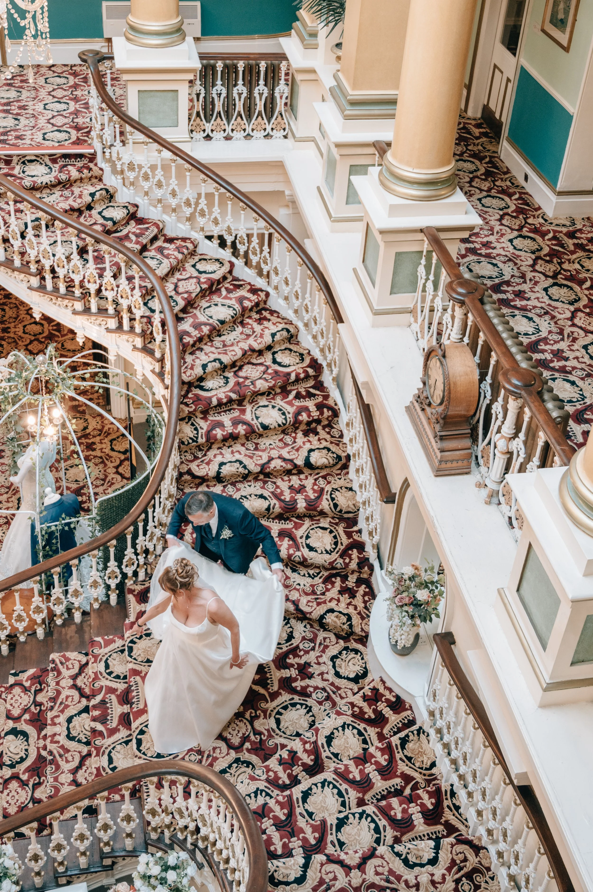 A bride and groom are descending a curved staircase with floral carpet at a wedding reception in a grand interior space with ornate columns, railings, and floral decorations.