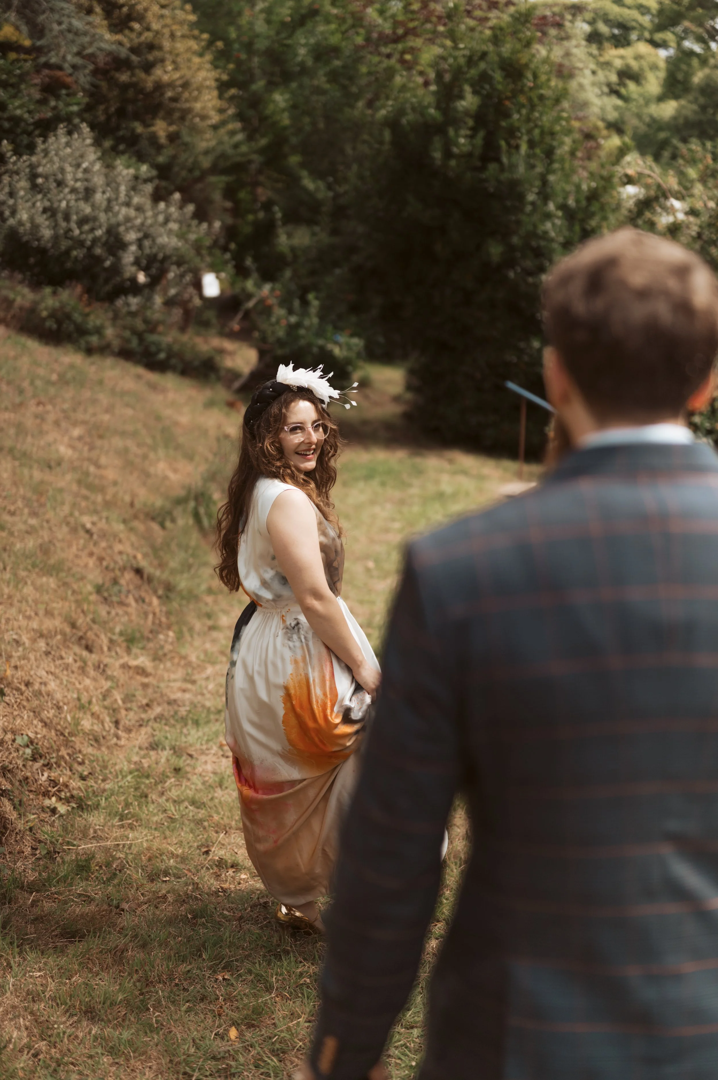 A woman with long, curly hair wearing a white dress with orange and brown patterns and a white fascinator, smiling at a man dressed in a dark plaid suit, outdoors in a park with trees and greenery.