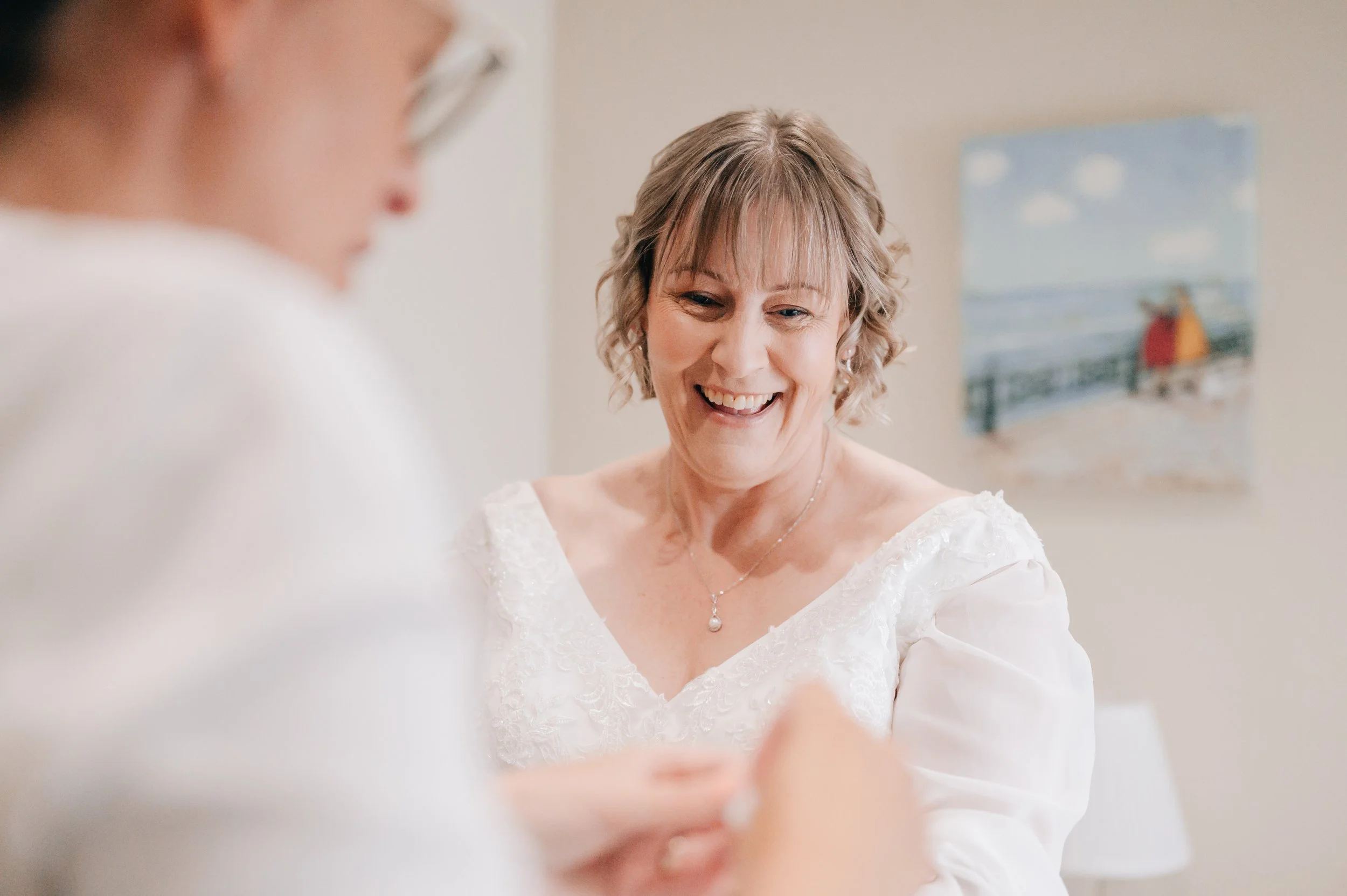 A smiling woman in a white dress with lace details, wearing a pearl necklace, is looking down at someone not visible in the image. A blurred person in white is in the foreground. A painting of a person in an orange coat on a bridge is on the wall in 