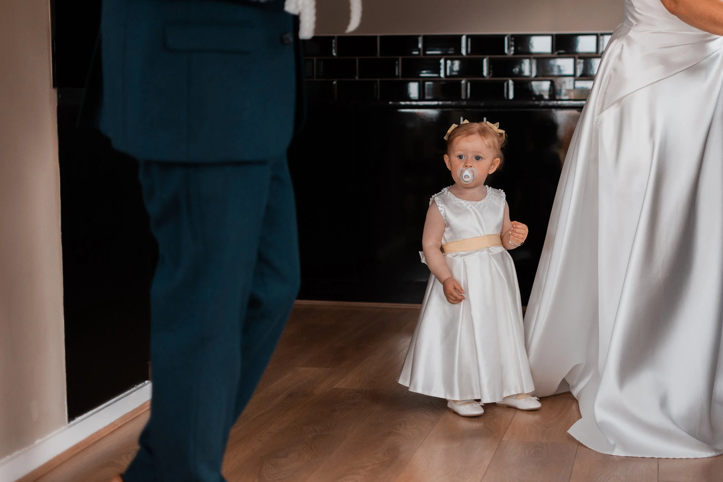A young girl in a white dress with a yellow sash and a pacifier in her mouth, standing on a wooden floor, looking at a person in a dark suit, with part of a white-draped table or curtain on the right side of the image.