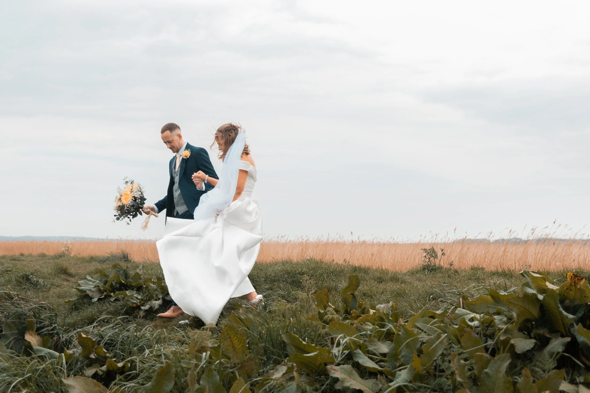 Wedding couple walking through a grassy field, the bride holding a bouquet of flowers, the groom in a suit, both smiling and holding hands.