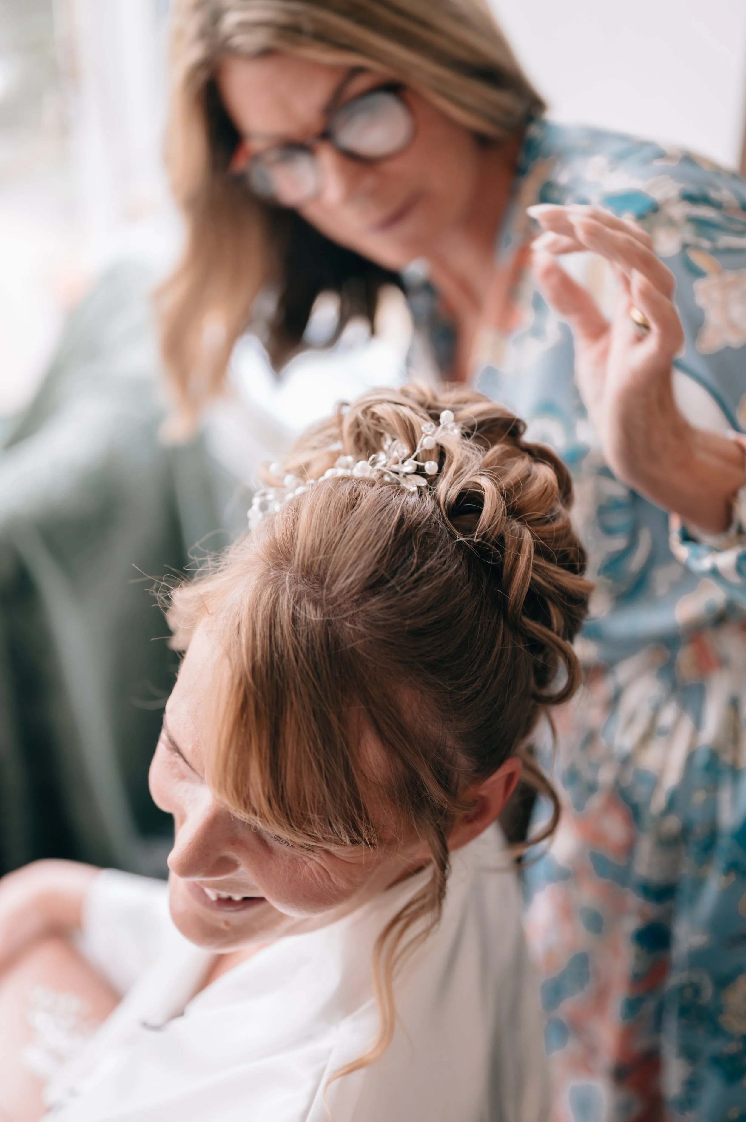 A woman with styled hair and hair accessories is smiling while another woman, possibly a hairstylist or family member, is adjusting her hair.