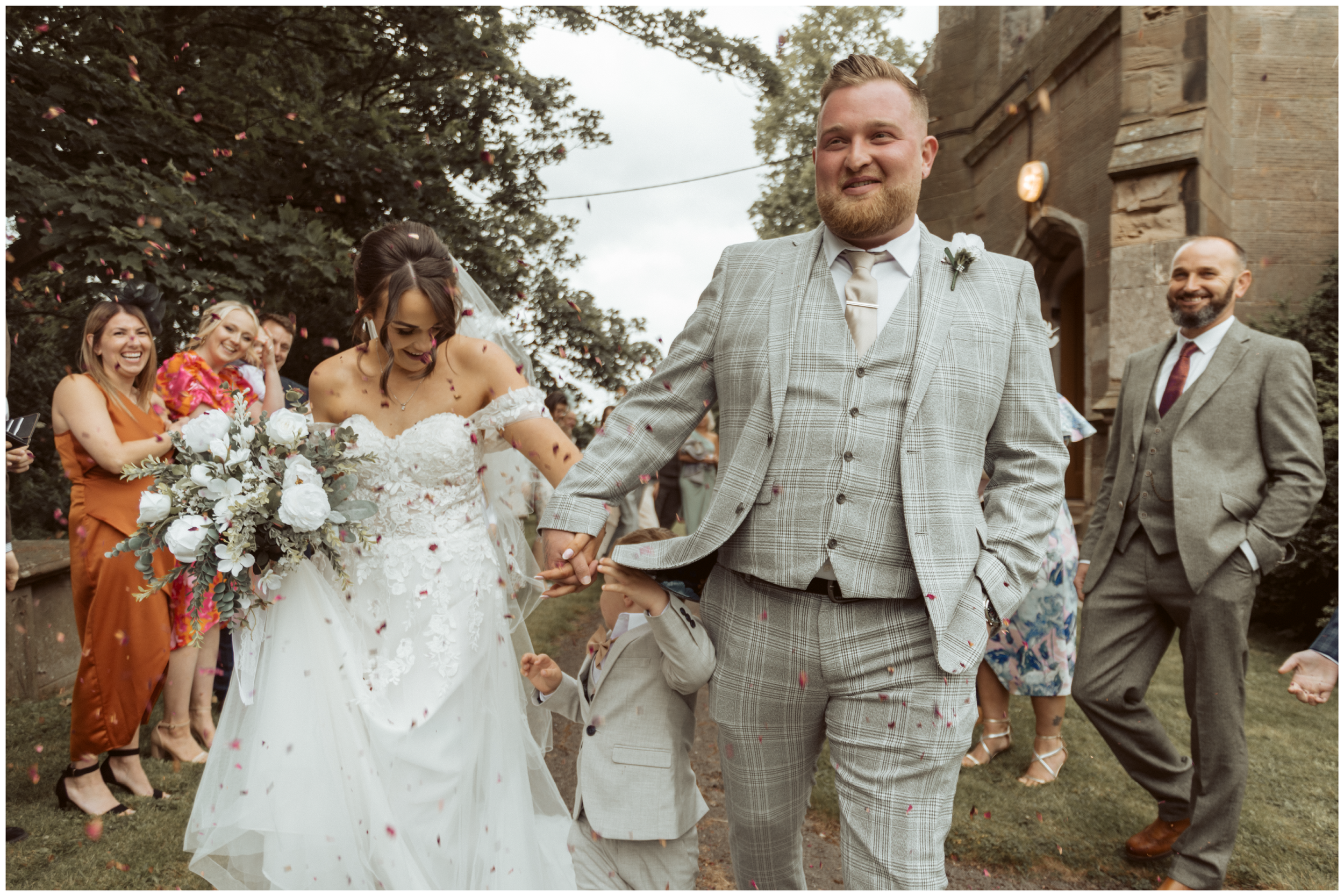 A bride and groom walking hand in hand at their wedding, surrounded by smiling guests outdoors with trees and a stone building in the background, as flower petals are thrown.