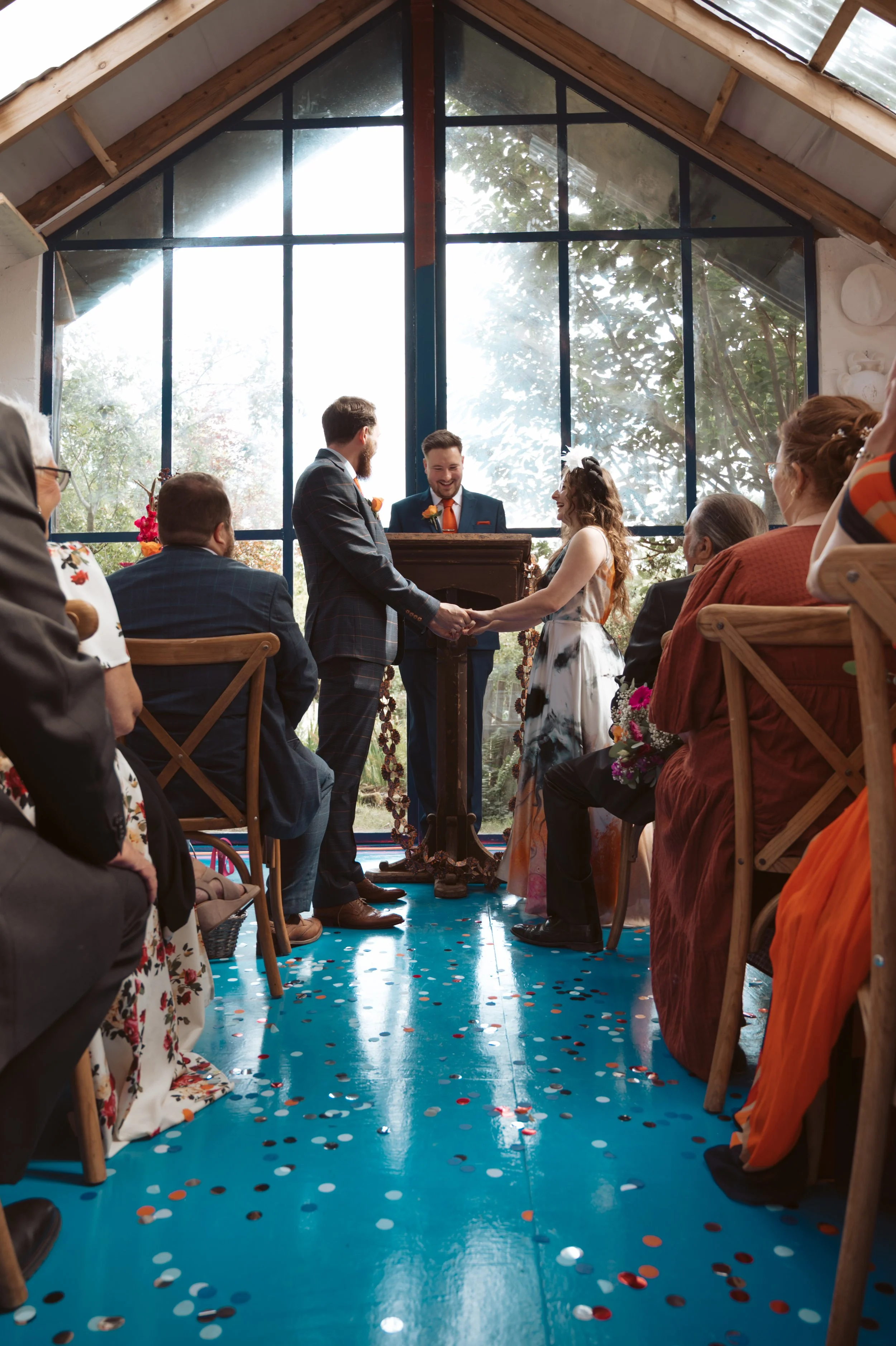 A couple getting married inside a glass-walled chapel, with the officiant and guests present. The couple is holding hands and exchanging vows, with a bright, tree-filled background visible through large windows. The floor is decorated with scattered 
