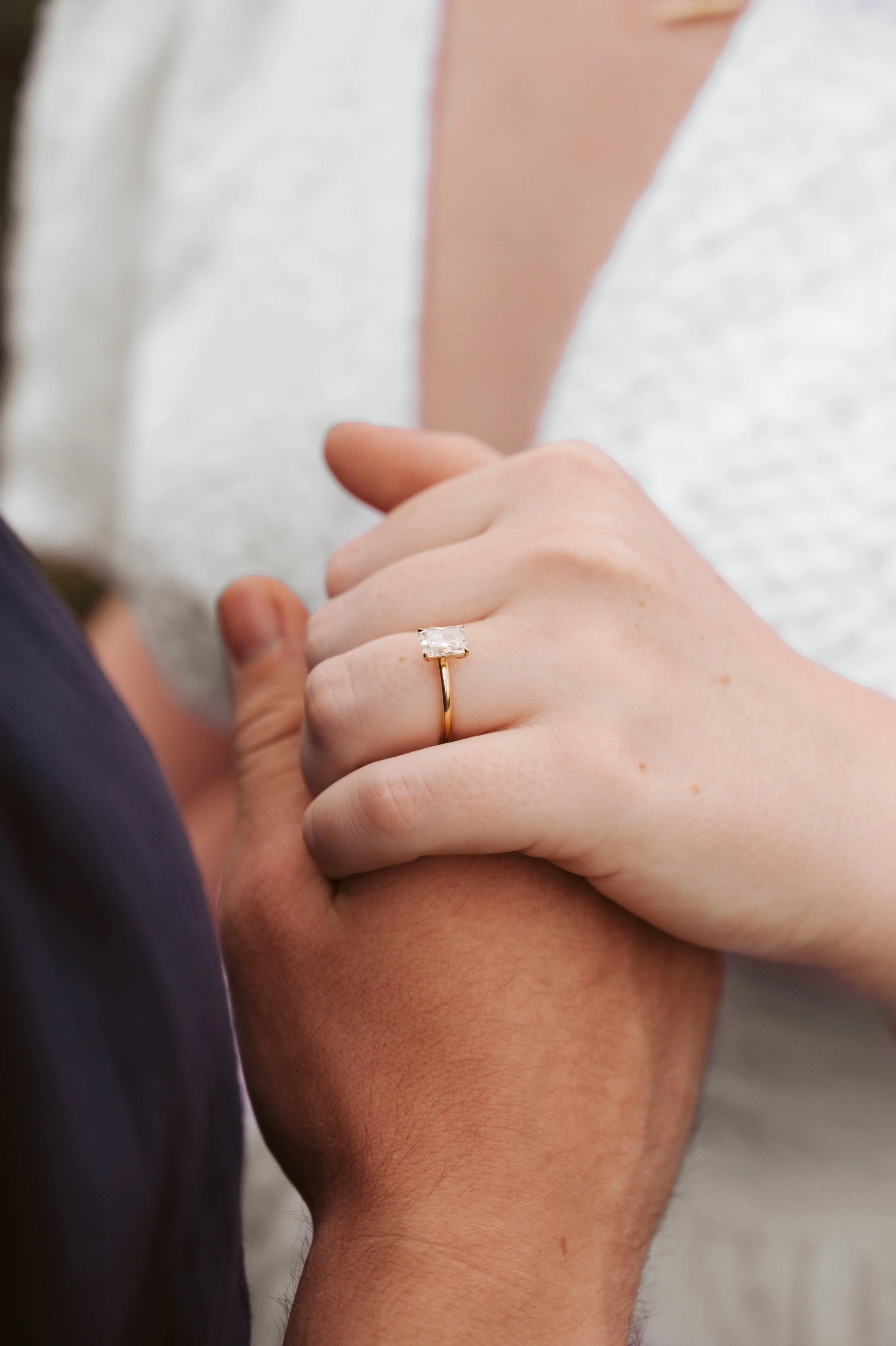 A woman wearing a white dress is holding the hand of a man with darker skin. The woman has a gold ring with a large rectangular diamond on her ring finger.