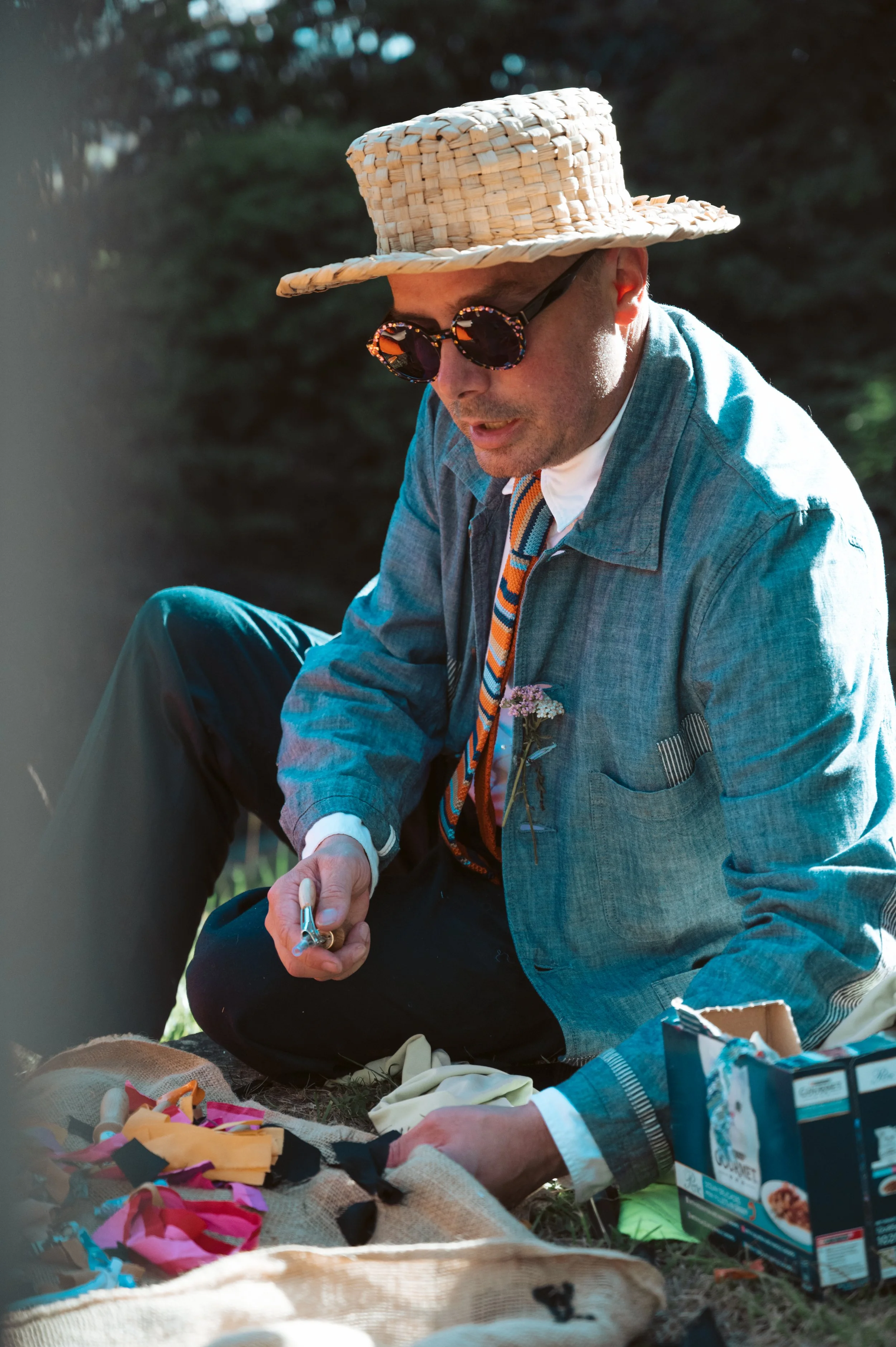 A man wearing a straw hat, sunglasses, a denim jacket, and a colorful tie sits outdoors with picnic items around him during daytime.