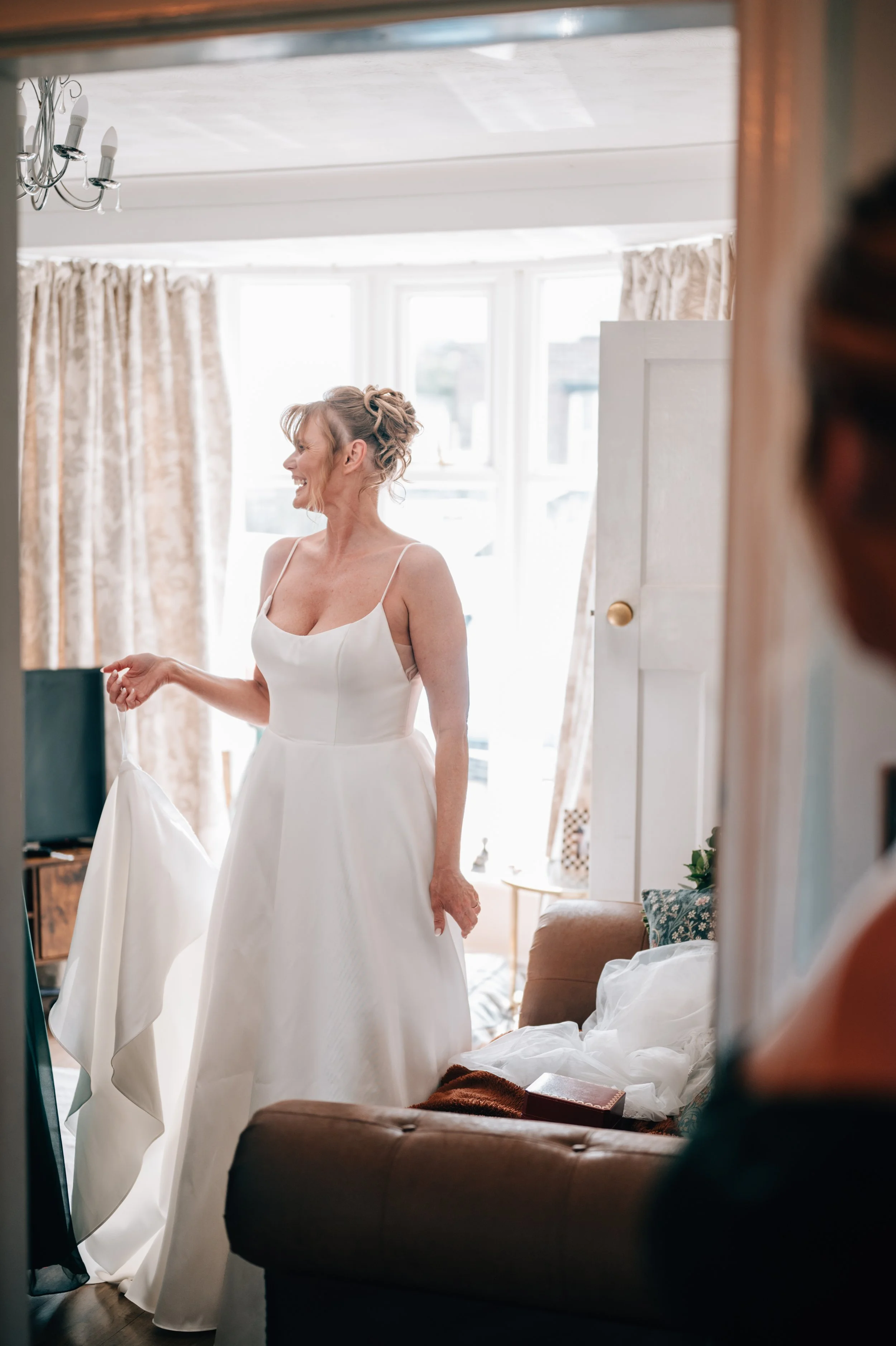 A woman in a white wedding dress smiling and holding her dress, standing in a sunlit living room with beige curtains and a brown couch.