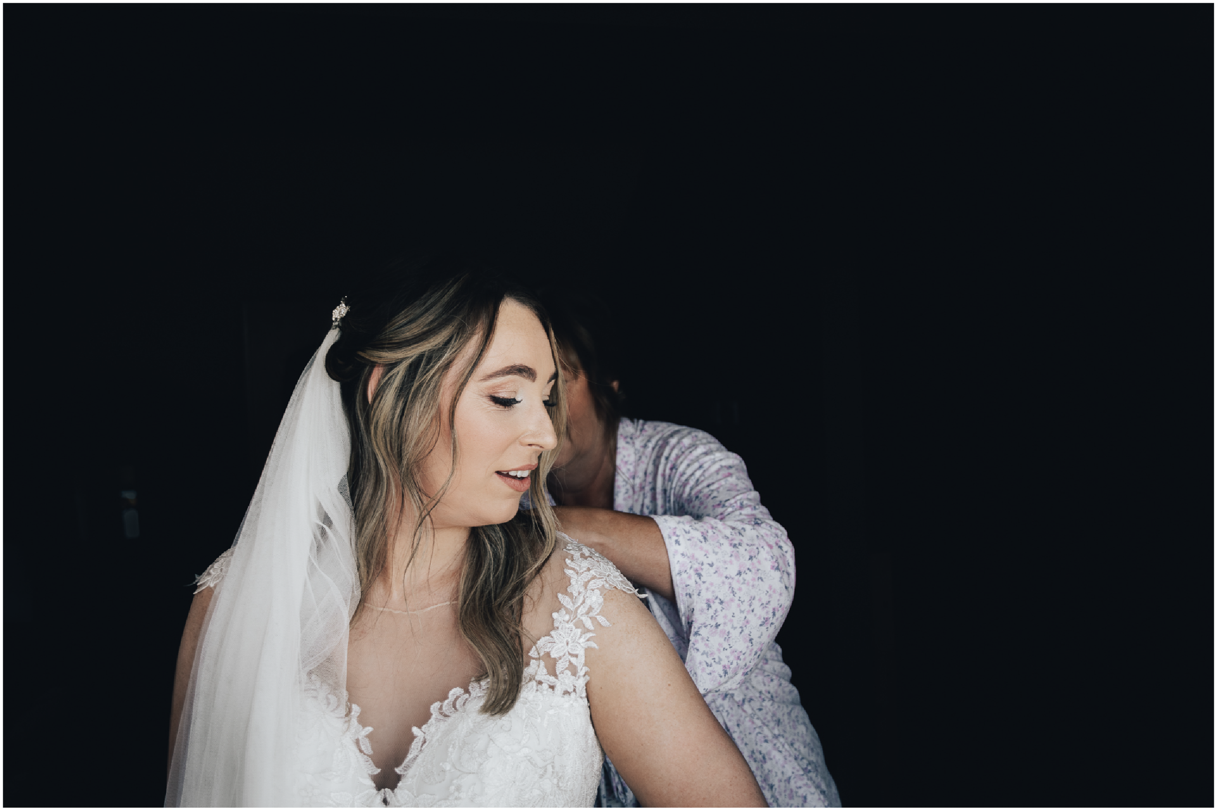 A bride with light brown hair in loose waves, wearing a white lace wedding dress and veil, is having her wedding dress adjusted by a person in a floral shirt.