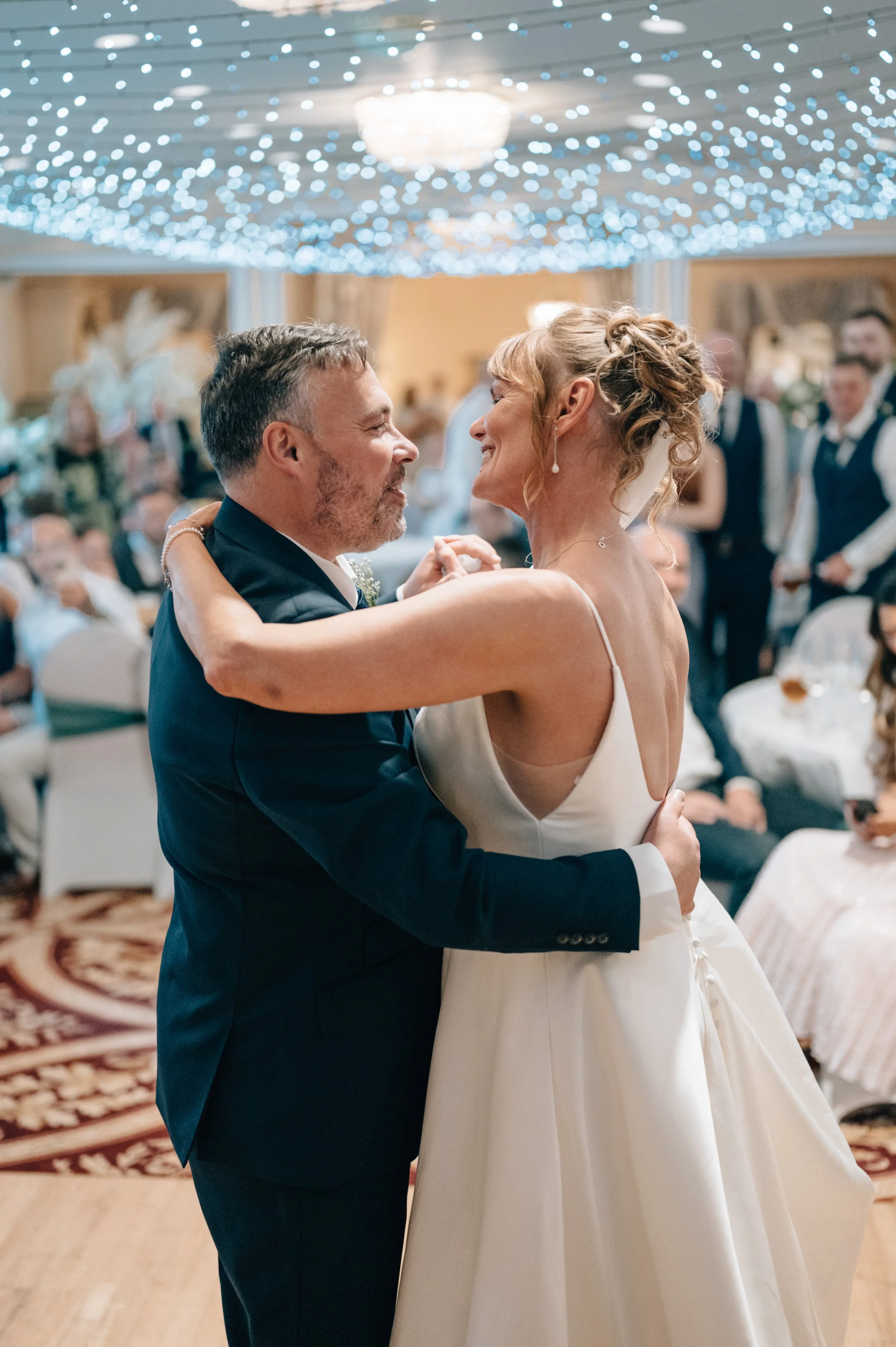 A bride and groom are dancing at their wedding reception, smiling at each other with guests watching in the background.