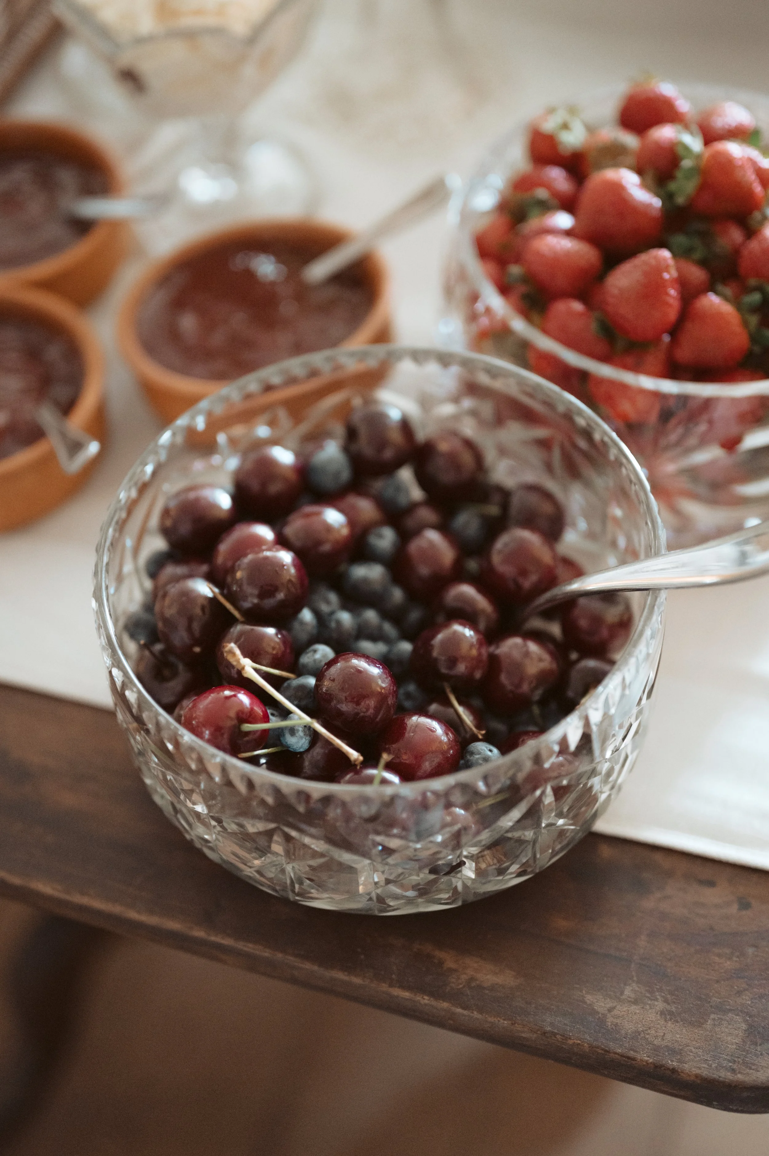Bowl of fresh cherries with a spoon, with bowls of strawberries and jam in the background on a table.