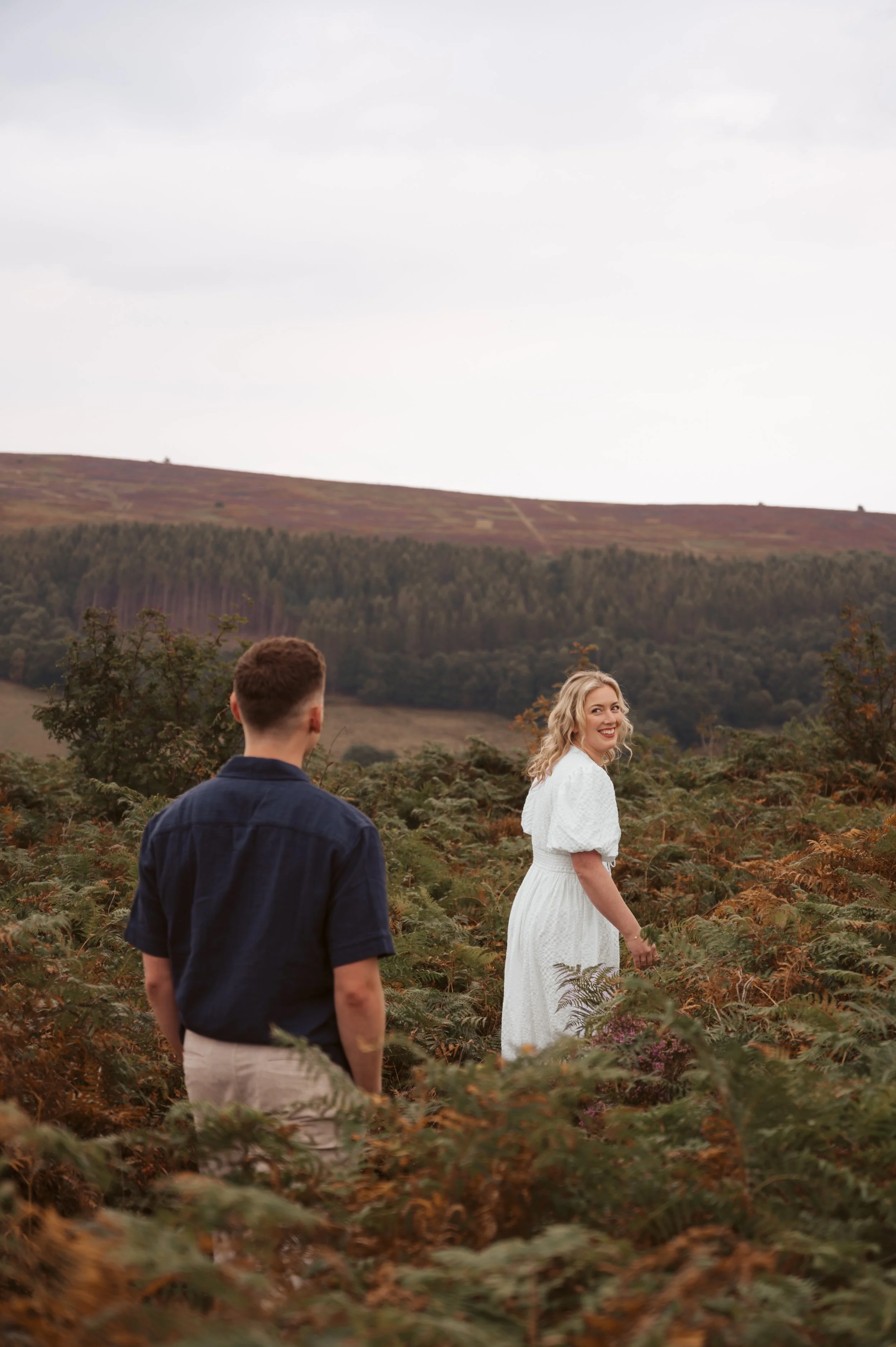 A smiling woman in a white dress walking through a field of ferns, with a man in a navy shirt and light pants standing nearby, in a rural landscape with rolling hills in the background.