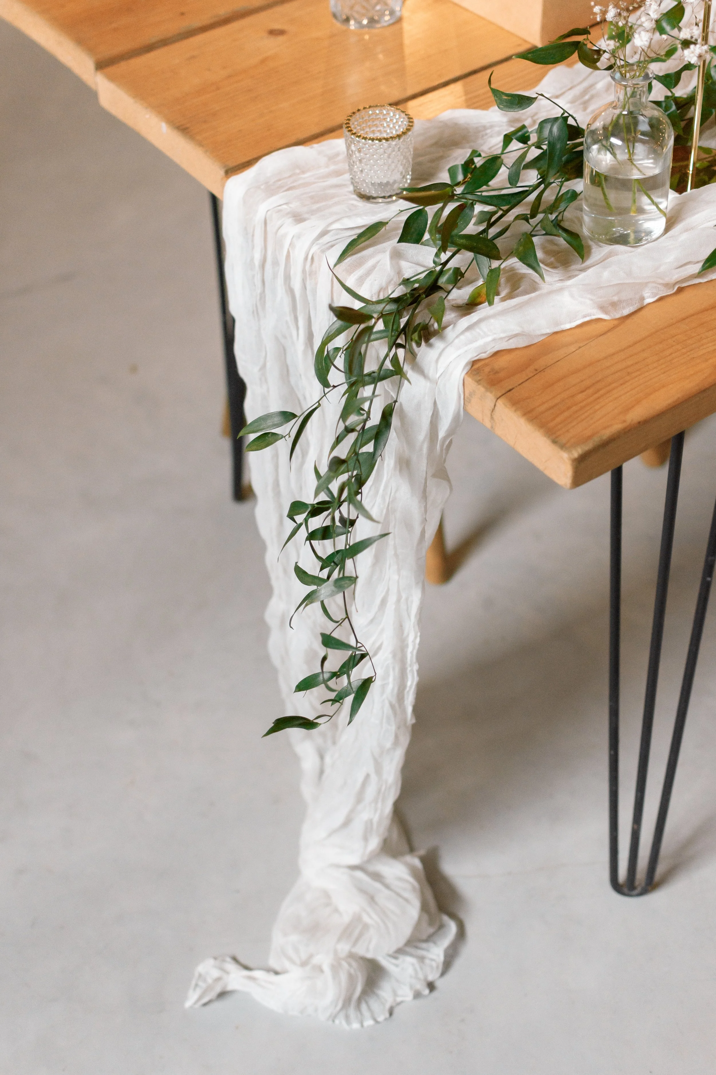 A wooden table decorated with a white cloth runner, glass vases with water and white flowers, a candle holder, and green leaves.
