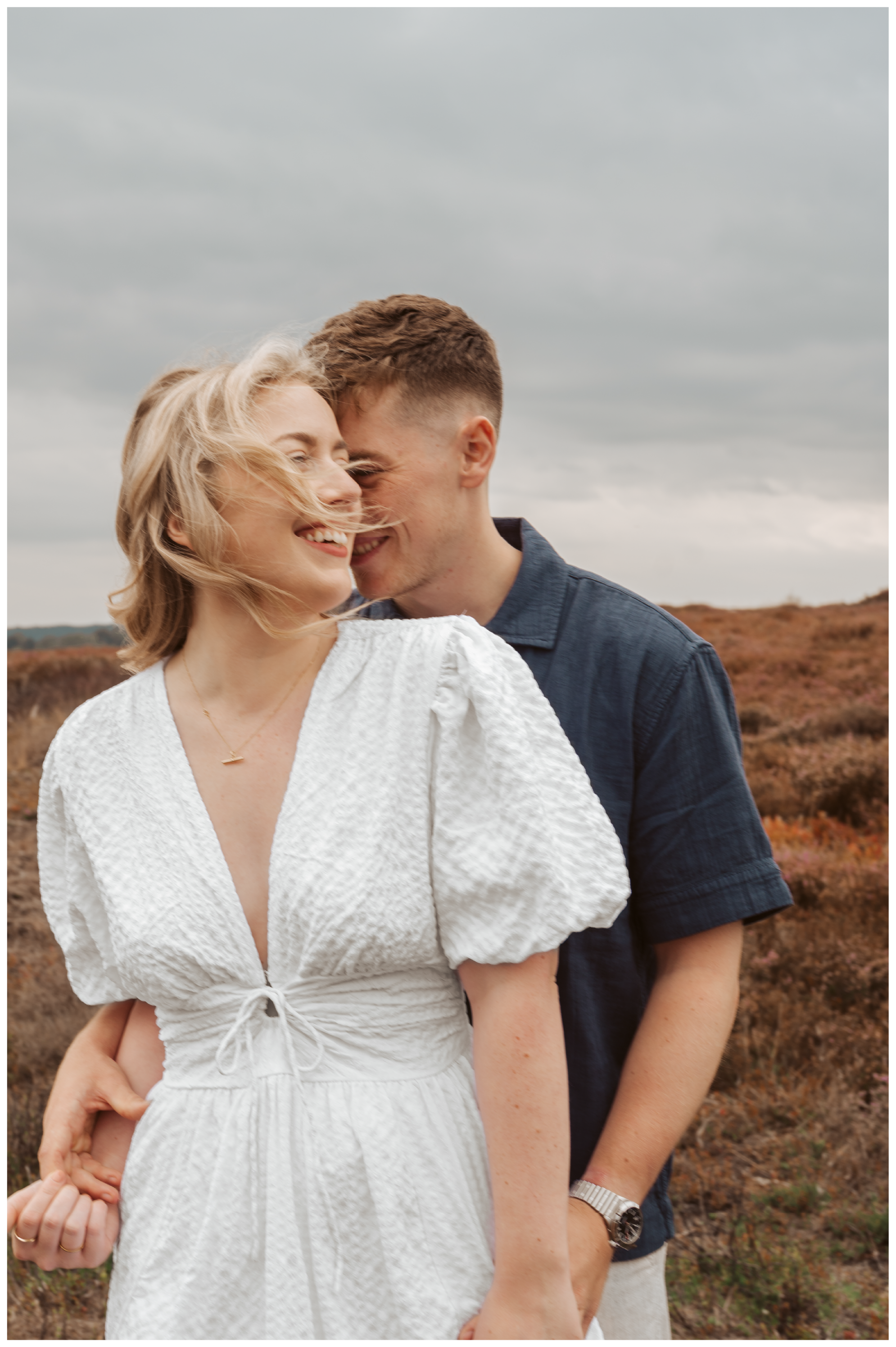 A couple smiling and close together outdoors, with overcast sky and brown shrubbery in the background.