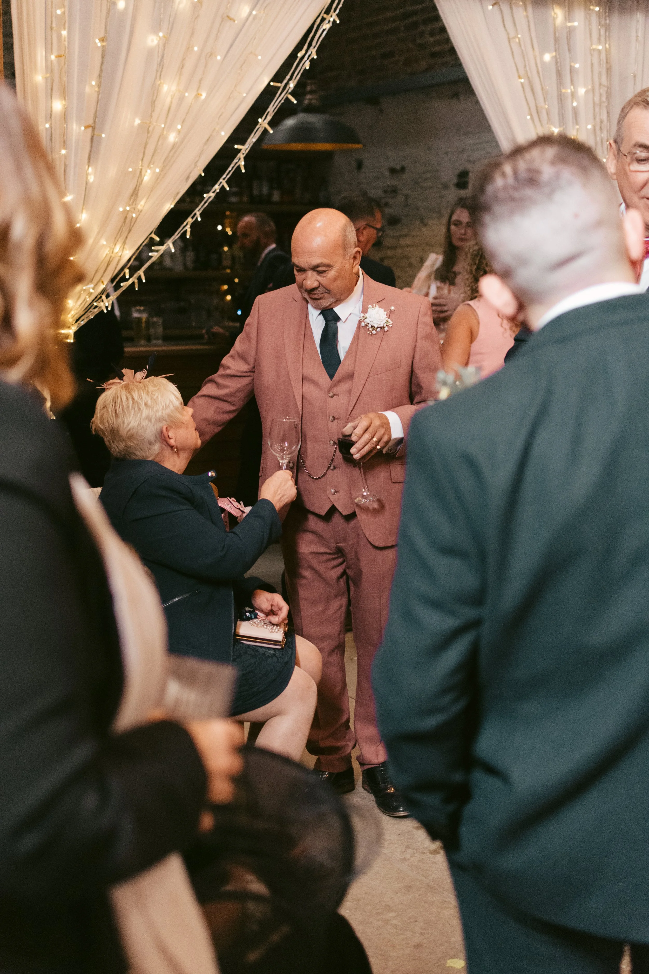 A man in a pink suit holding a wine glass stands in a crowd at a party, with a woman sitting and holding a champagne flute. The setting is decorated with string lights and a draped curtain.