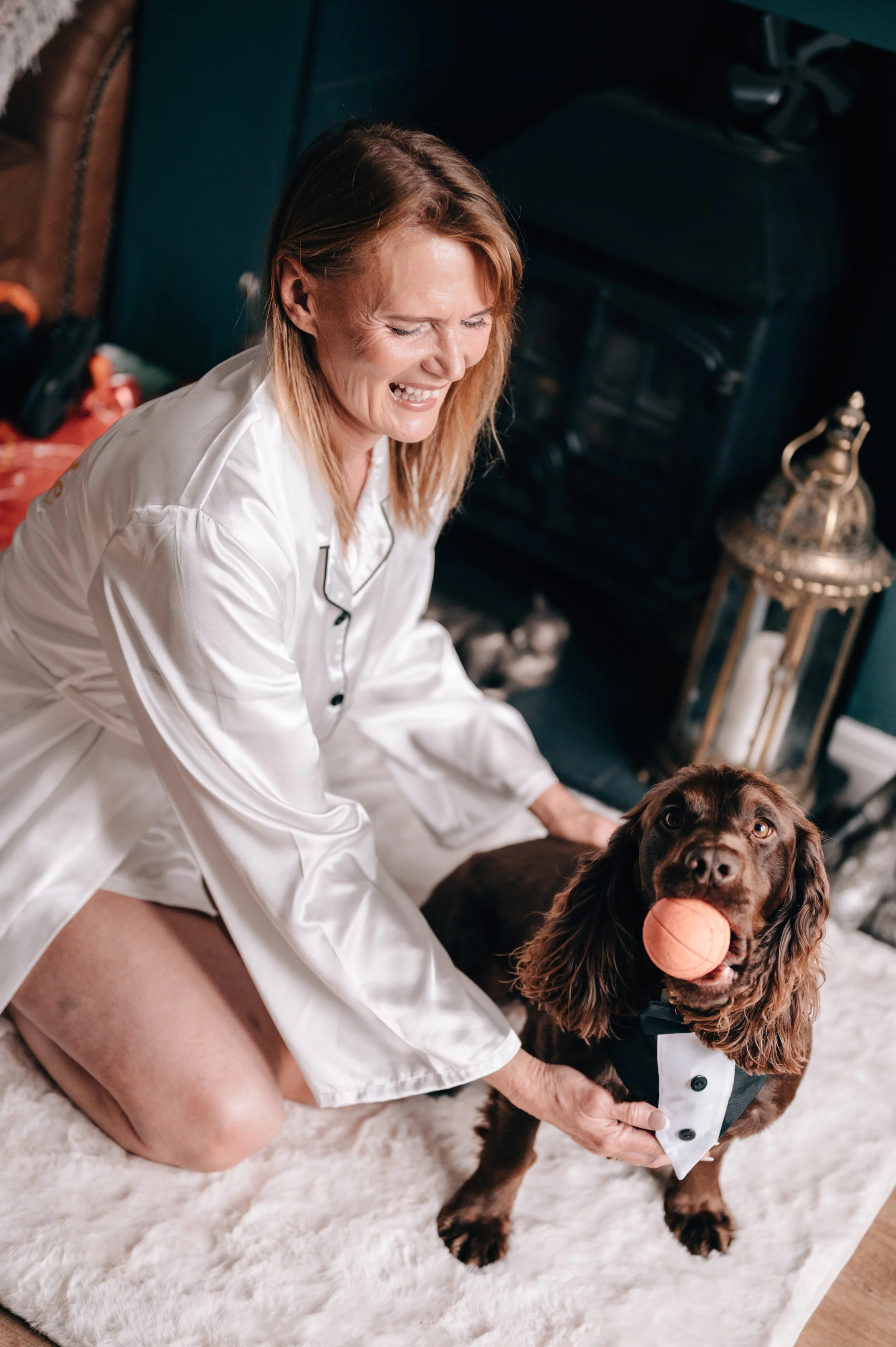 A woman in white pajamas kneels on a plush rug, smiling while holding a brown dog with a ball in its mouth. The dog is wearing a tuxedo.
