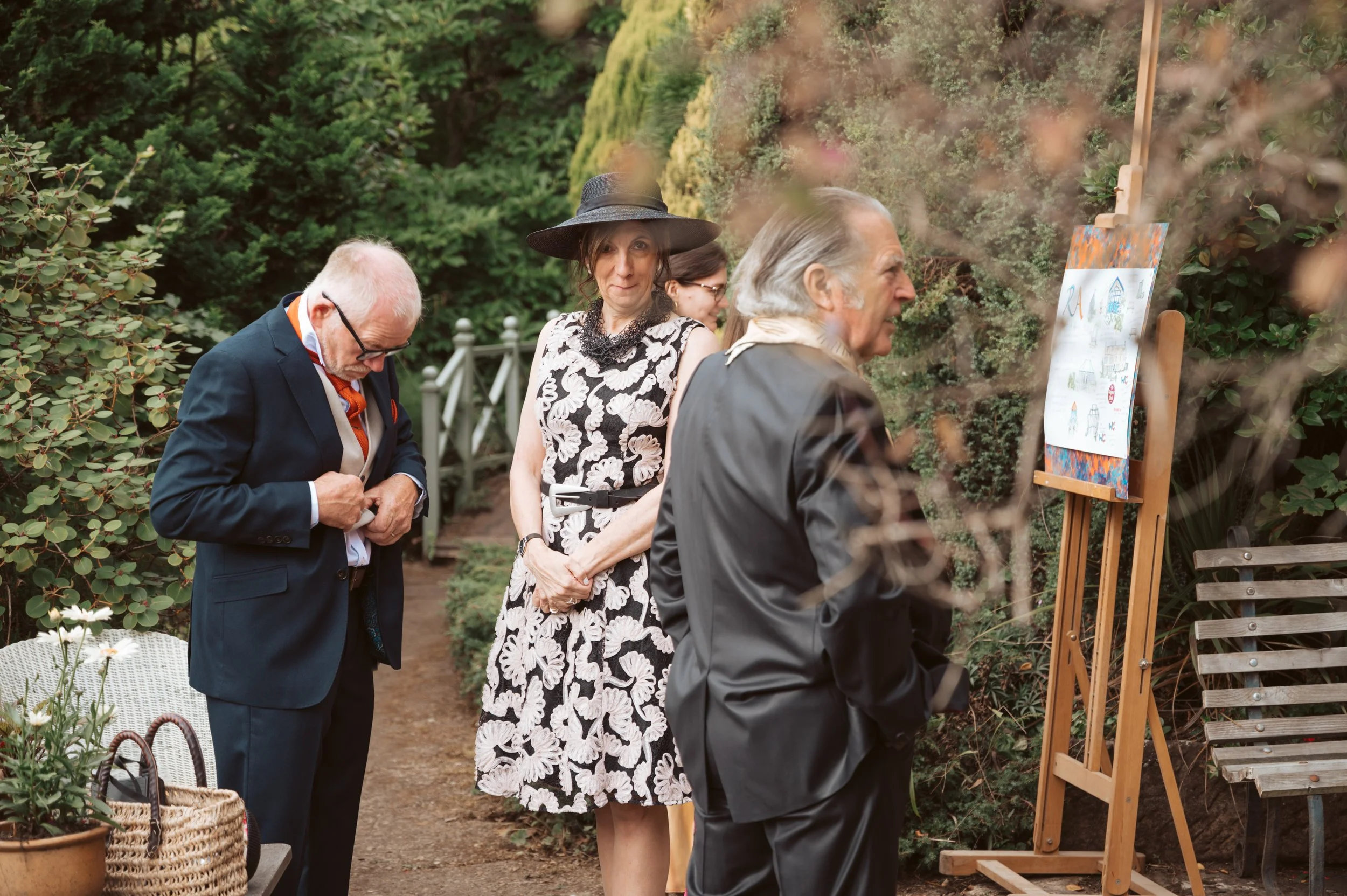 People viewing artwork on an easel outdoors in a garden, dressed in formal attire.