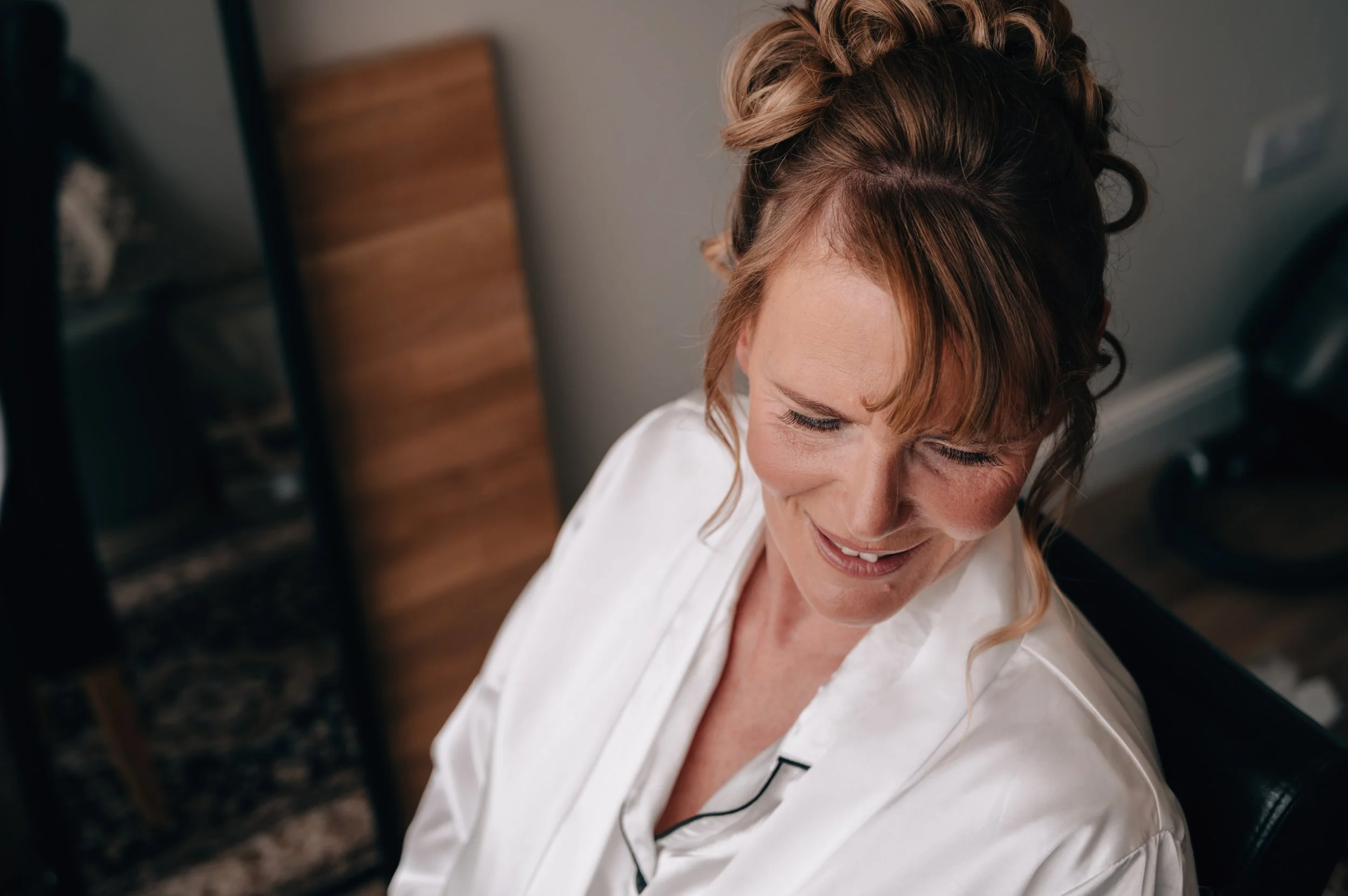 A woman with reddish hair styled in loose curls, smiling with closed eyes, wearing a white satin robe, likely getting ready for a special occasion.