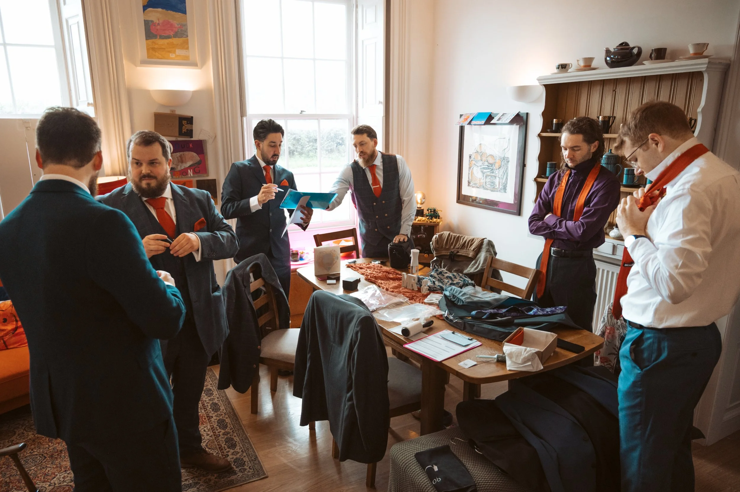 Group of six men in suits and ties preparing for an event in a well-lit room with a large wooden table, chairs, and various clothing and accessories on the table and chairs.