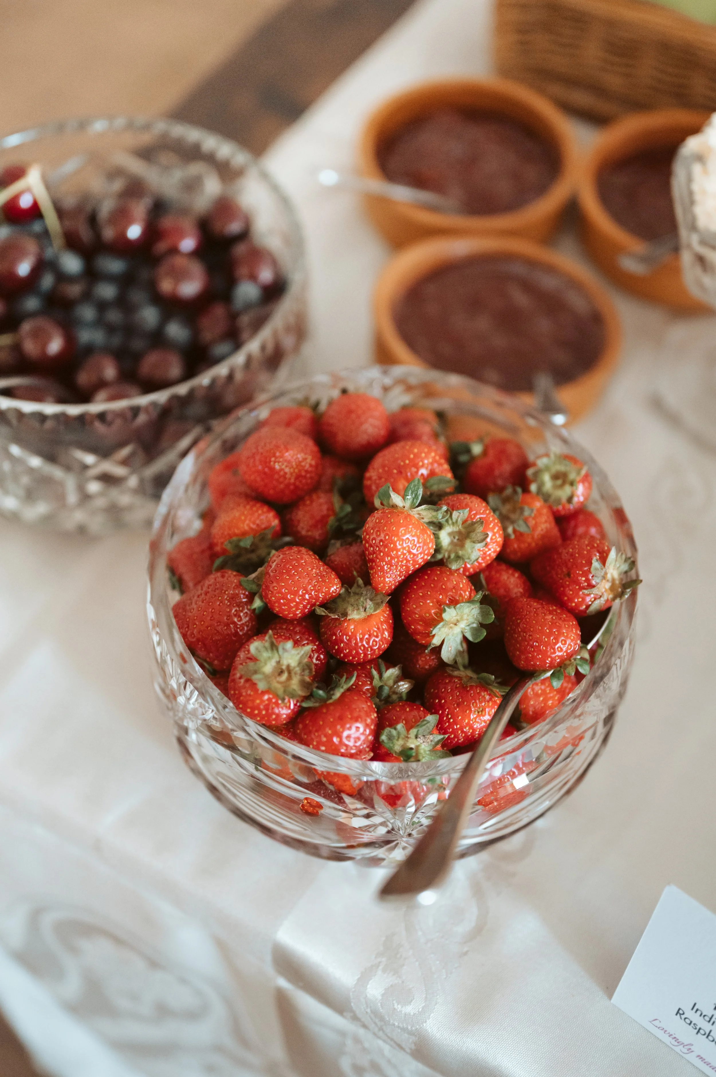 A glass bowl filled with fresh strawberries on a table, with bowls of jam and other bowls in the background.