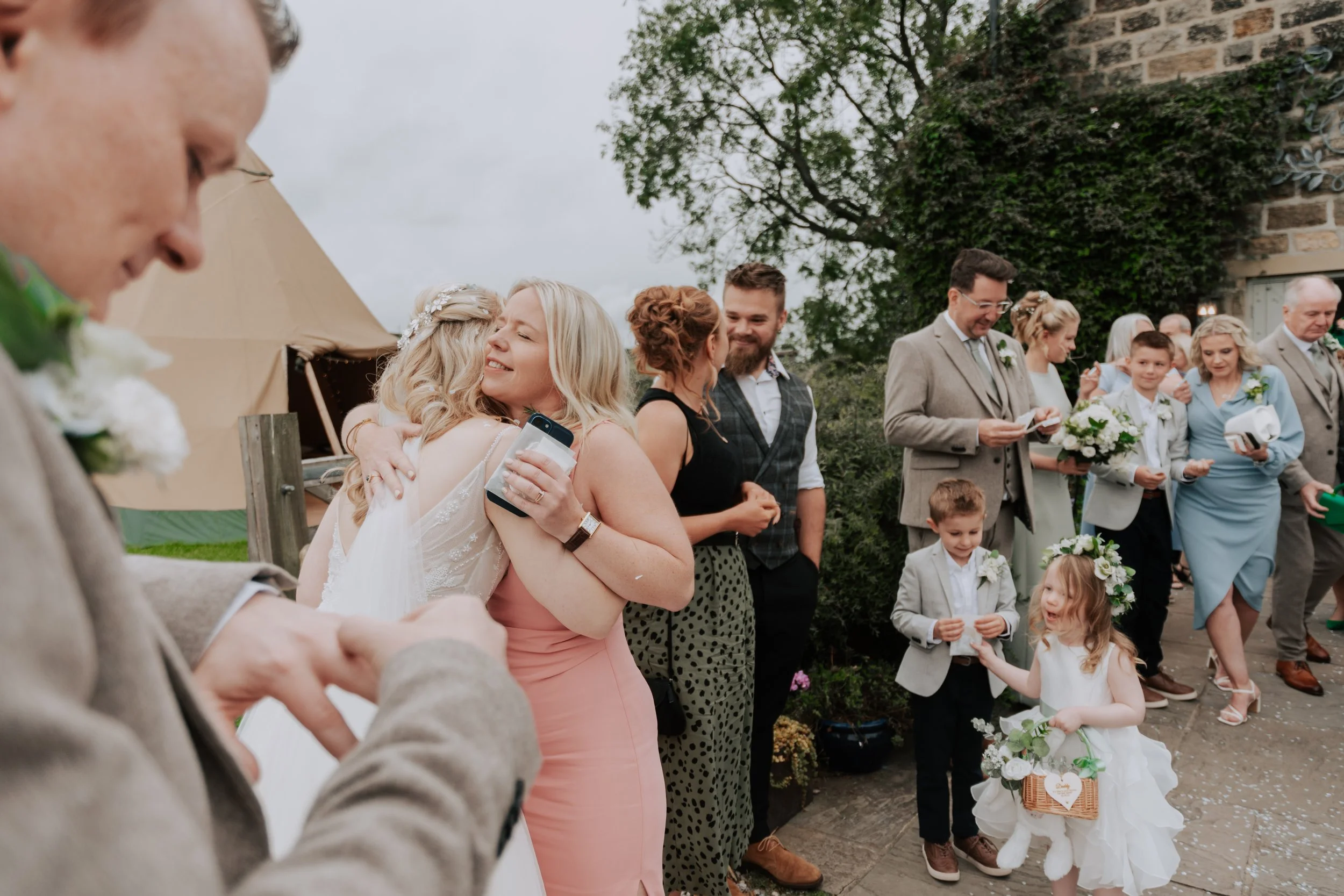 People celebrating at a wedding, some embracing, others holding flowers, and children dressed in formal attire, outdoors near a brick building and trees.