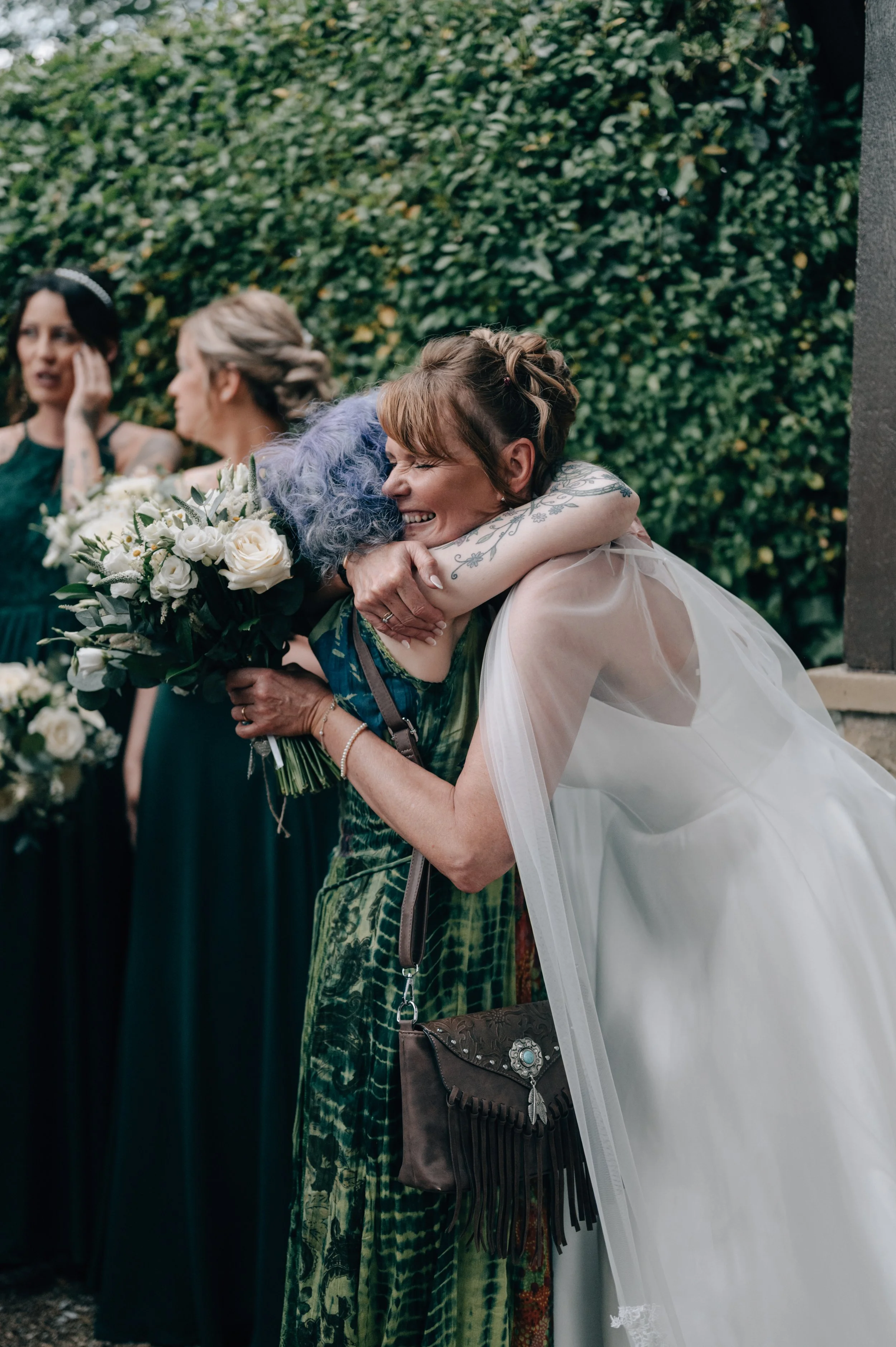 A woman in a white wedding dress is hugging an older woman with purple hair, who is holding a bouquet of white roses, at a wedding celebration.