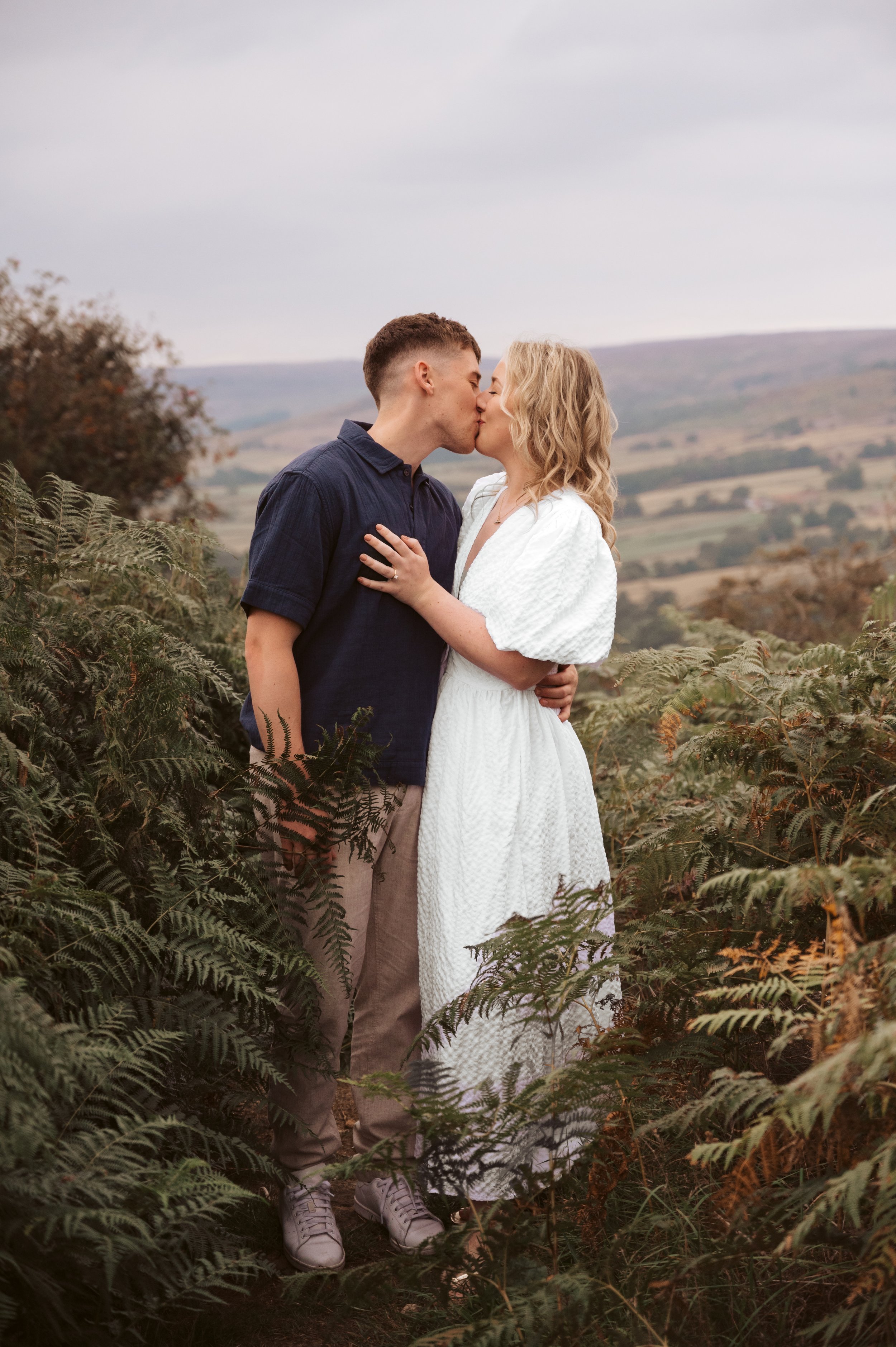 A couple sharing a kiss in a natural outdoor setting with hills and greenery.