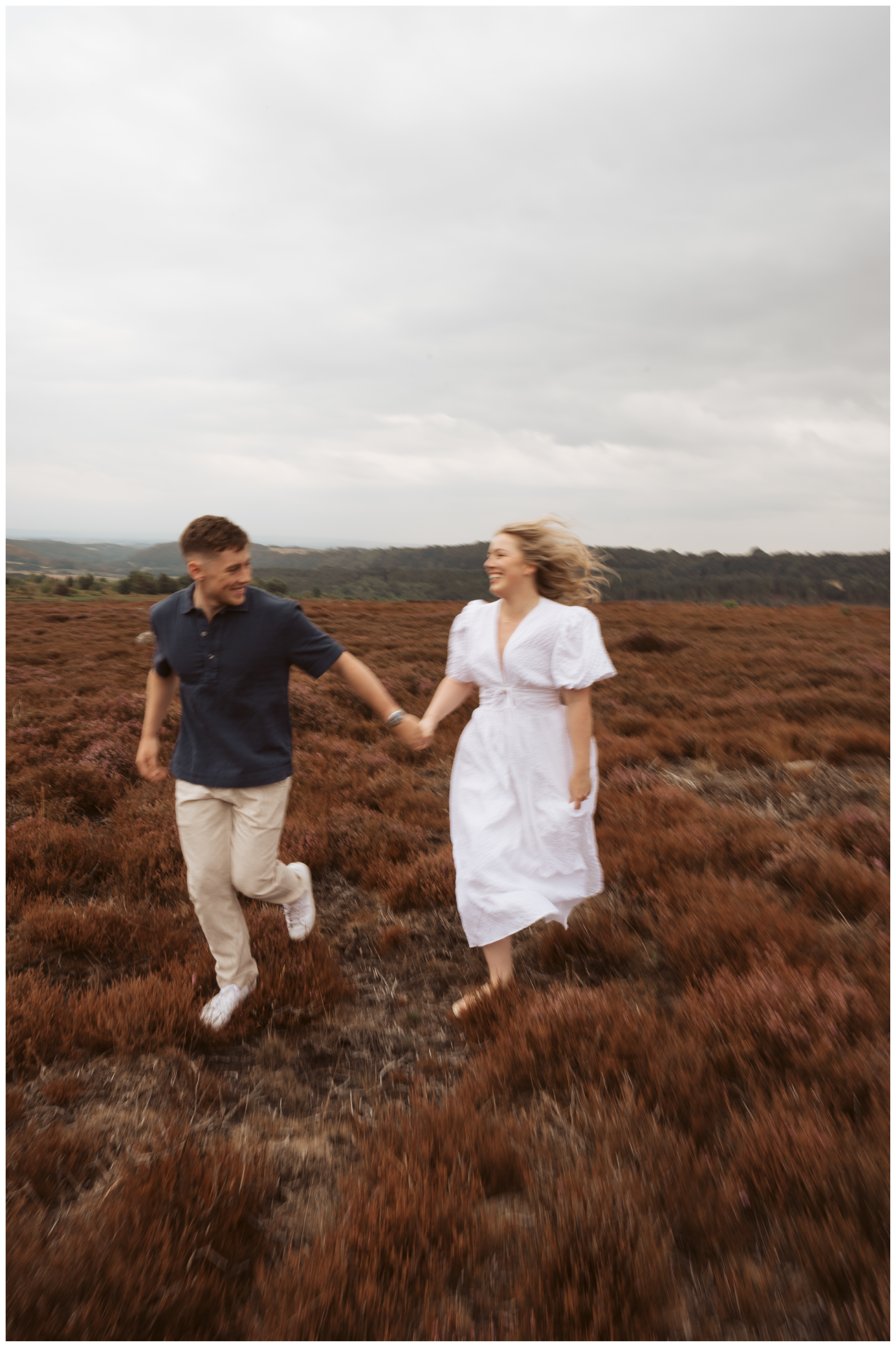 A couple holding hands and running outdoors on a dirt path through a field of reddish-brush, with overcast skies.