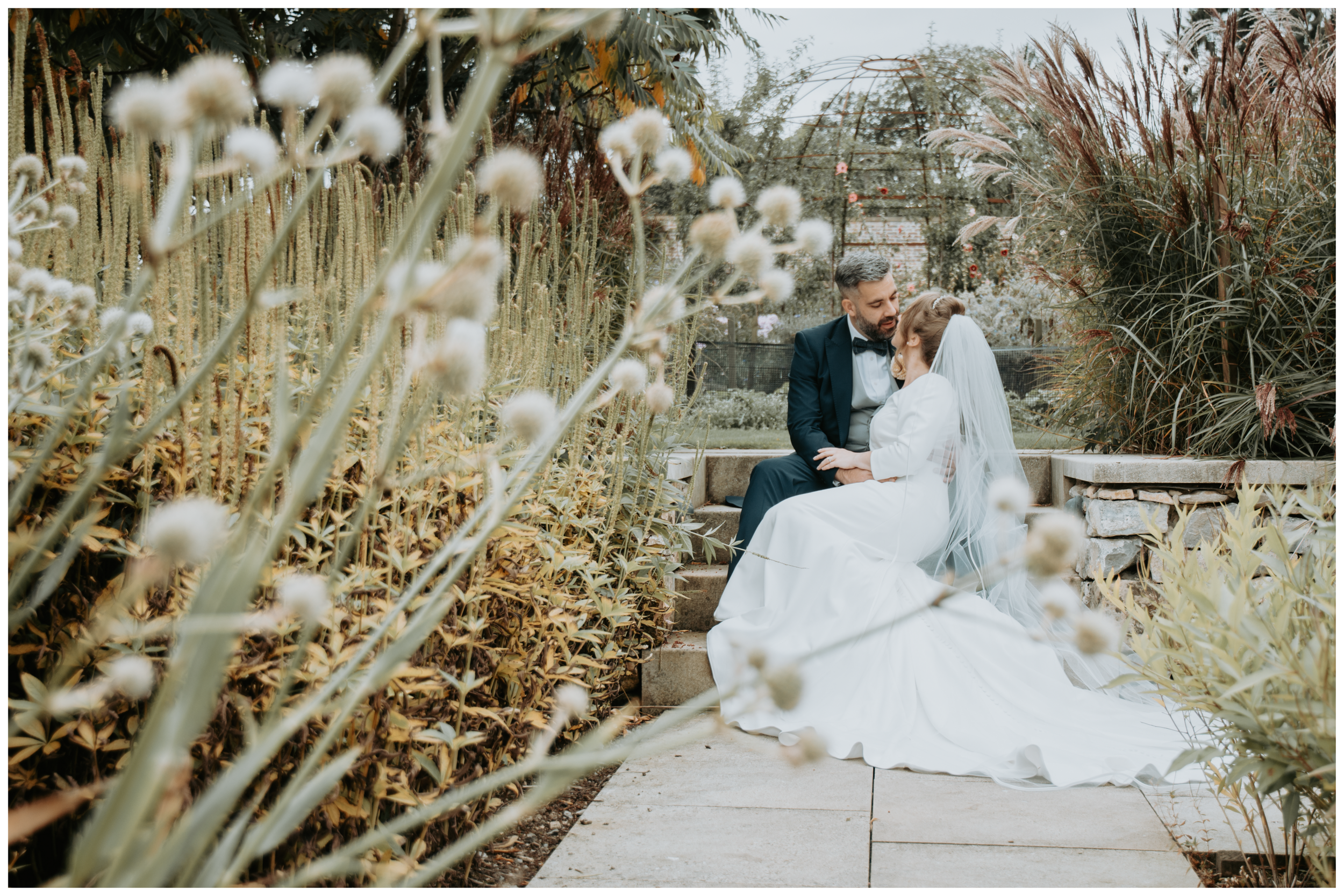 A wedding couple sitting on stone steps in a garden surrounded by tall plants and flowers, with the bride in a white gown and veil and the groom in a tuxedo. The couple looks at each other affectionately.
