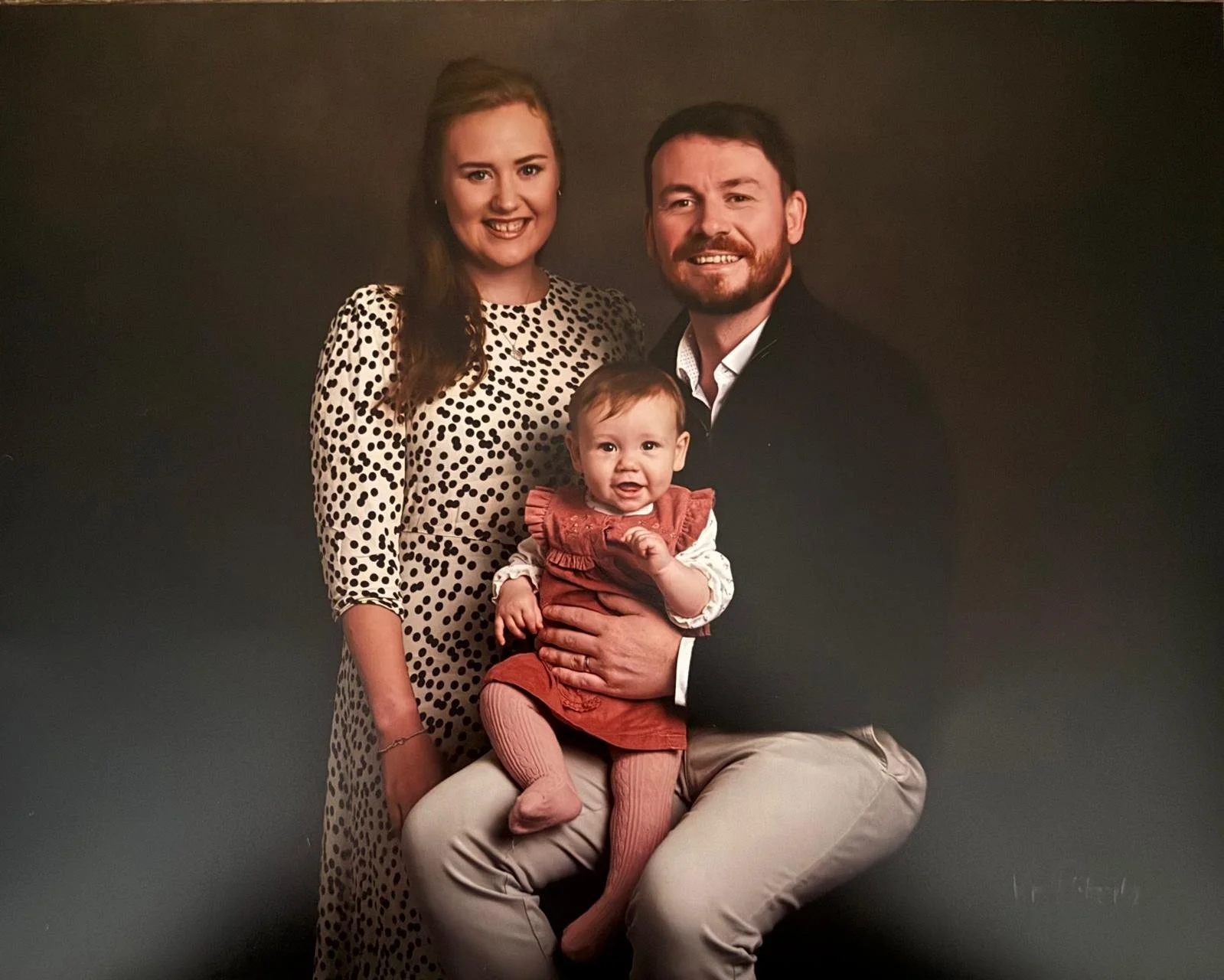 Family portrait of a woman, man, and a baby girl against a dark background. The woman is wearing a polka dot dress, and the man is dressed in a suit with a white shirt. The baby girl is sitting on the man's lap, wearing a ruffled rust-colored dress and pink tights, with all three smiling.