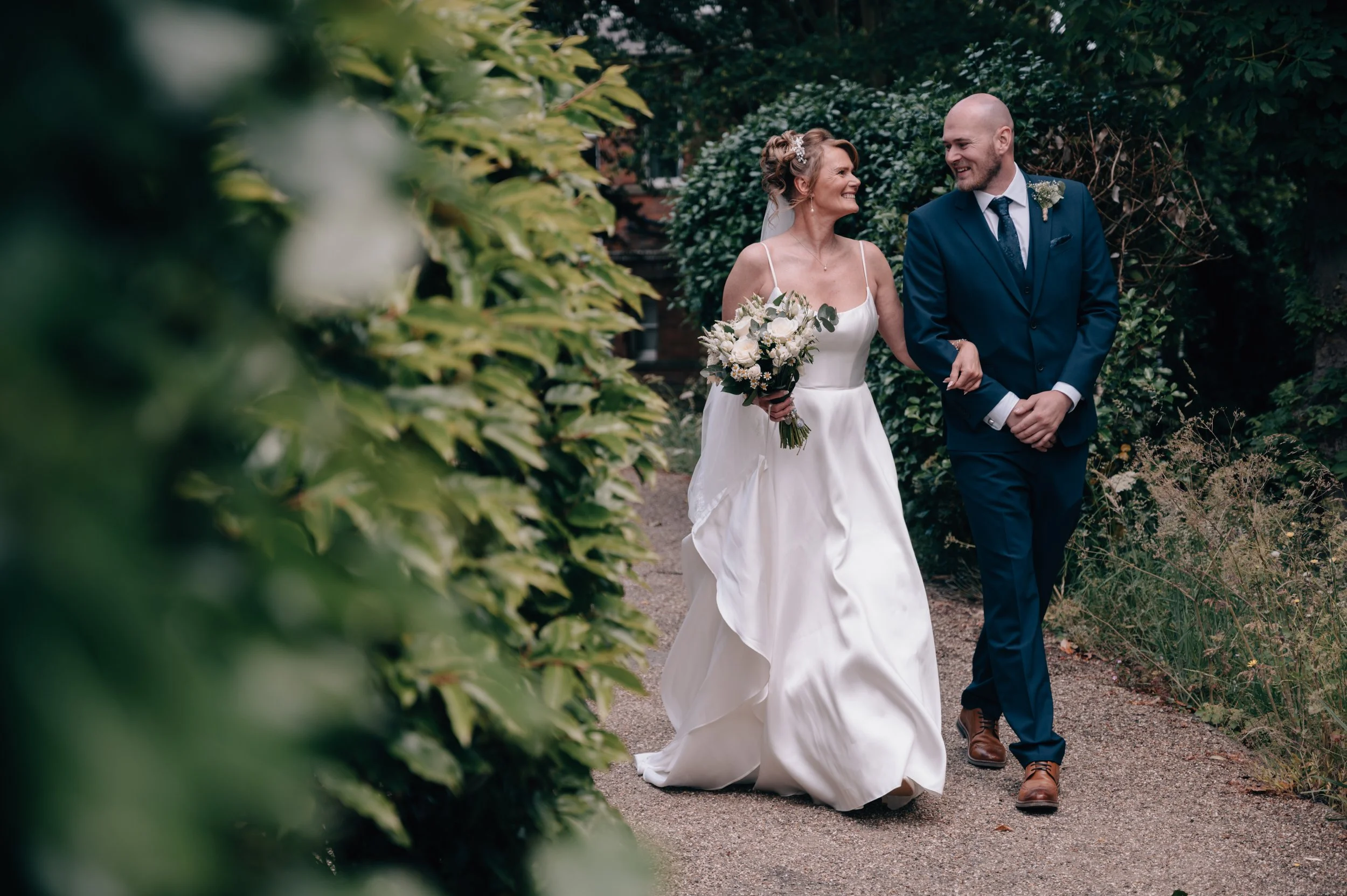 A bride and groom walking arm in arm on a garden path, smiling at each other on their wedding day. The bride is wearing a white wedding gown and holding a bouquet of flowers, while the groom is dressed in a navy suit with a tie and boutonniere.